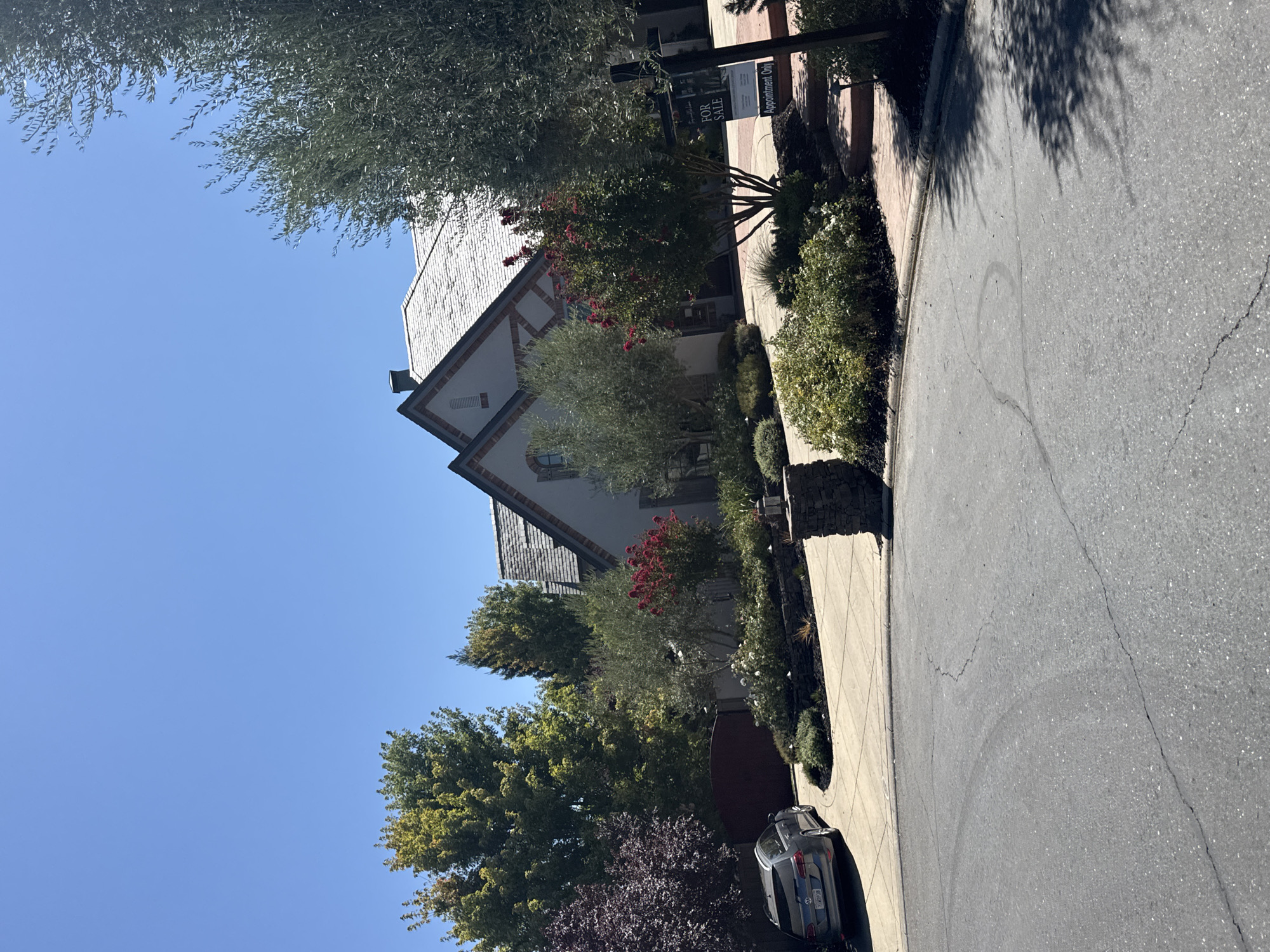 This is a photograph of a residential property with a "For Sale" sign and a car on a sunny day. The house, a multi-story structure with a gray tiled roof, is partially visible behind trees and landscaping. The front yard features various plants, including flowering bushes and trees, and a paved driveway. A dark-colored station wagon is parked on the driveway. A stone pillar with a light fixture stands near the entrance to the property. The scene is captured from a street that appears to be a residential road with cracked asphalt. The sky is clear and blue. The image does not contain any people. The location is identified as Roseville, United States.