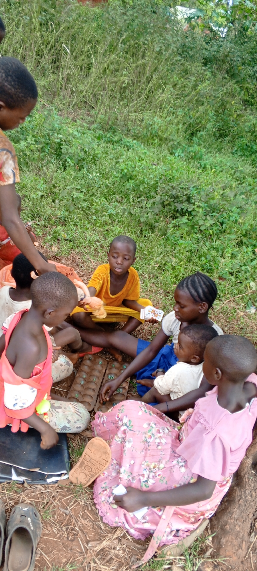 In Ngalula, Democratic Republic of the Congo, a group of at least seven children of varying ages are gathered outdoors on the ground. The setting features a mix of reddish-brown dirt and sparse dry grass where they are sitting, with a dense background of lush green vegetation suggesting a natural or rural environment. The scene is illuminated by daylight, indicating a clear or bright day.
The children are engaged in various forms of play and interaction. At the center, two wooden Mancala boards, filled with green playing pieces, are laid out, with some children actively involved in the game. A girl in a yellow outfit holds a doll and some playing cards, looking forward. Another child in a red top is engrossed in the board game. A girl with braided hair, wearing a white top and blue skirt, cradles an infant on her lap while observing the game. To her right, a girl in a pink floral dress and matching top sits, seemingly relaxed. Other children are partially visible, participating in the group activity.
Notable details include simple clothing, typical of daily life. A brown flip-flop with "GUCCI" written on its sole is visible near the girl in the pink dress. Black sandals or crocs are also on the ground in the foreground. The children appear focused and calm, absorbed in their activities. The overall impression is one of communal play and interaction among children in a natural, outdoor setting.