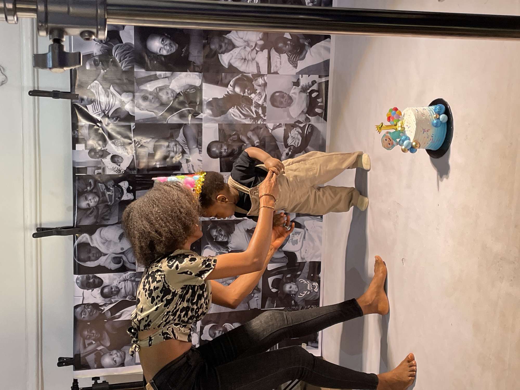 In a photography studio, a barefoot woman with curly hair, wearing a patterned cropped top and black jeans, is holding up a small child in tan overalls and a golden crown. The child faces a birthday cake decorated with blue and white frosting, colorful balloons, and a "CoComelon" character, which is attached to a white wall. Behind the woman and child is a backdrop featuring a collage of black and white photos of people, secured by clips on a metal frame. The background wall is a solid, light color. The location is implied to be Jos, Nigeria.