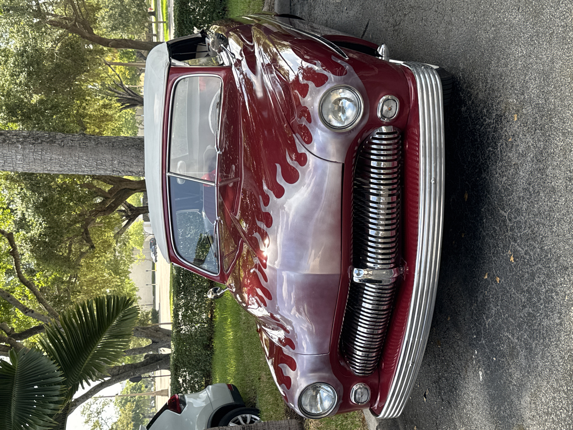 A burgundy 1951 Mercury convertible with silver flames is parked on asphalt in front of a manicured hedge and grass. In the background, trees with green foliage and a light-colored building are visible. To the left of the Mercury, the rear of a white SUV is partially visible. The scene appears to be in Coral Springs, Florida, United States of America.