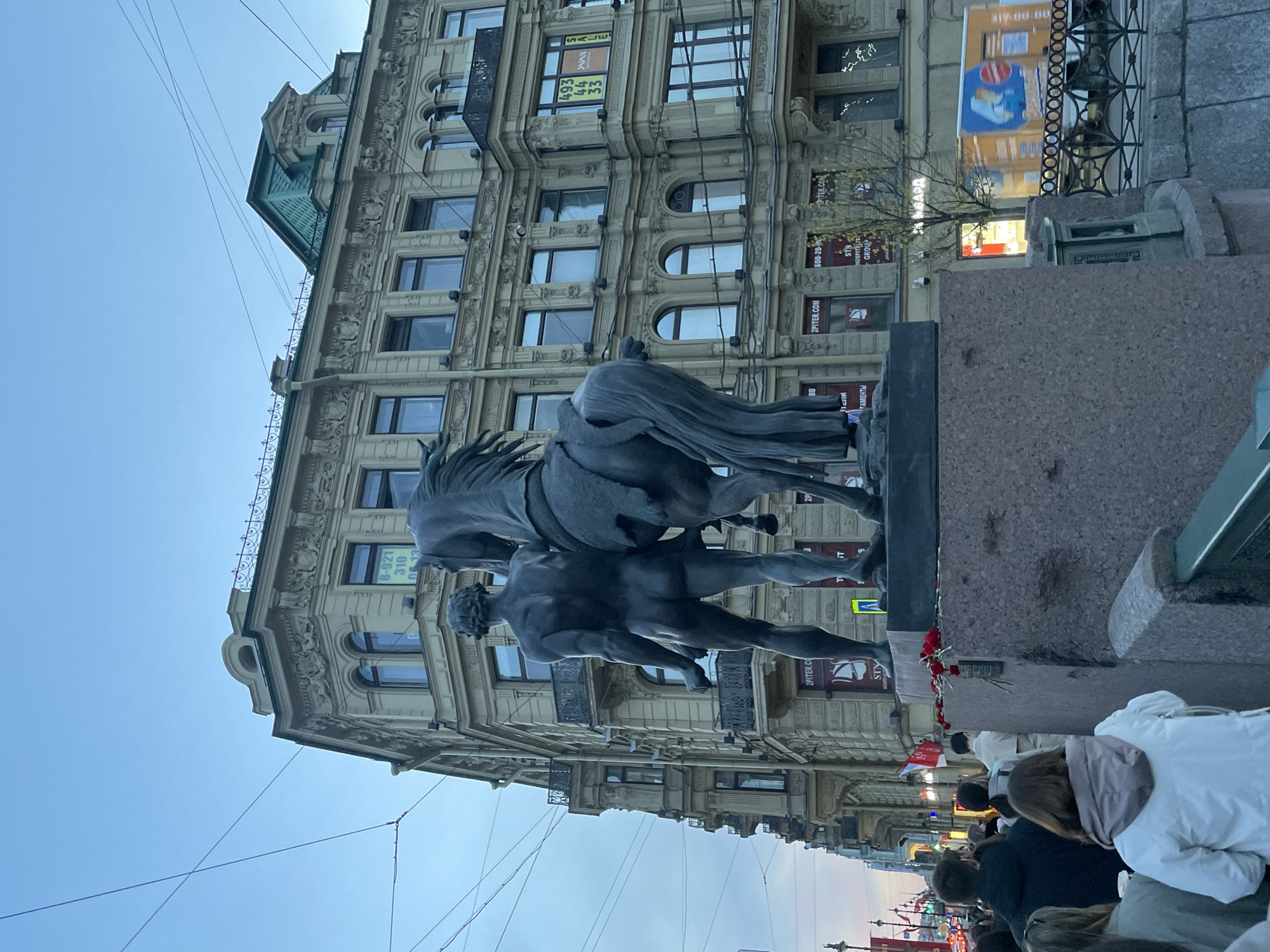 The image shows a statue of a man wrestling a horse in Saint Petersburg, Russia. The statue is mounted on a stone pedestal. Behind the statue is a large, multi-story building with ornate architectural details and numerous windows. There are commercial advertisements and signage visible on the building's facade. A crowd of people is gathered at the base of the statue. The sky is visible in the upper portion of the image, appearing slightly overcast.