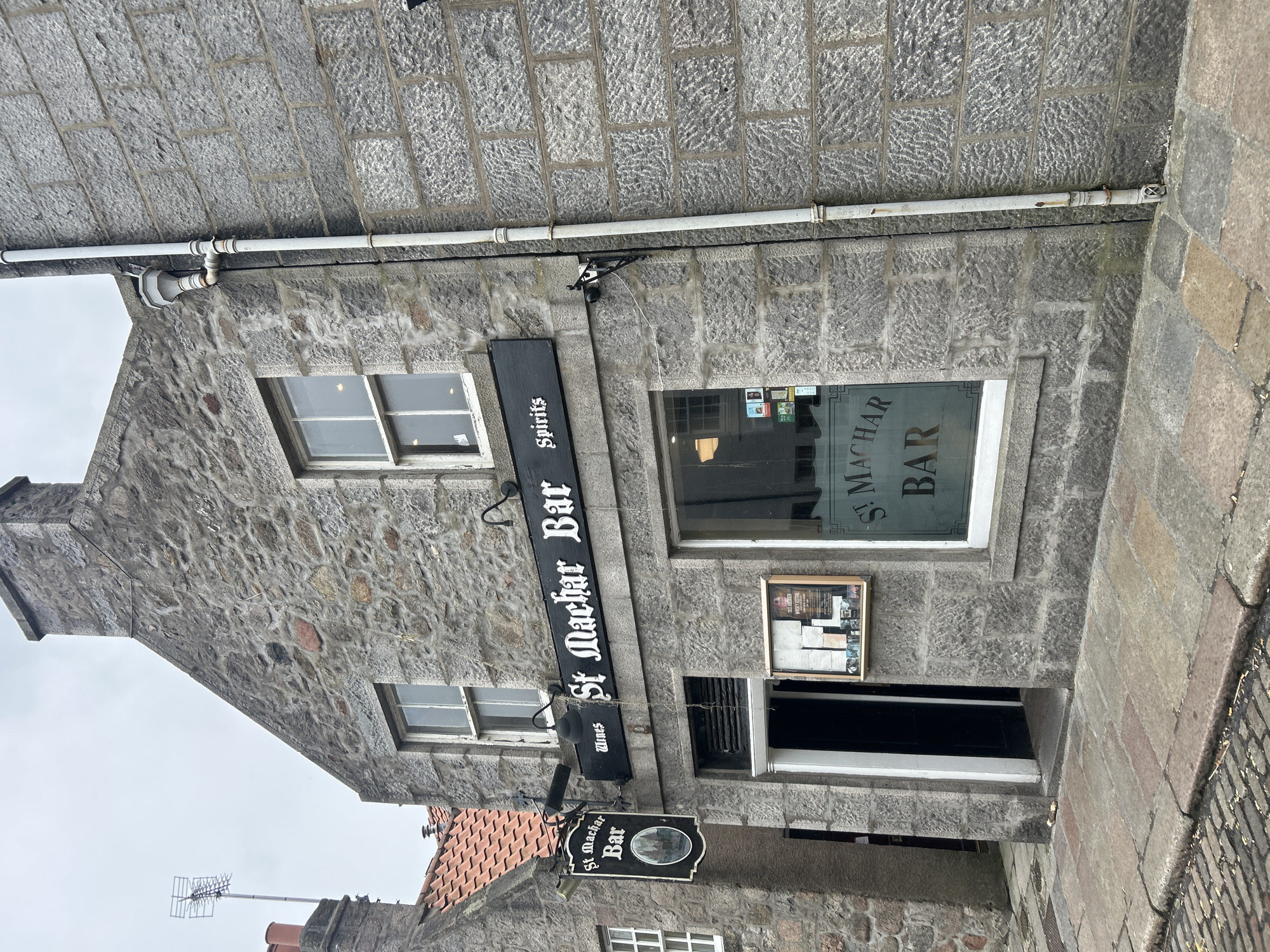 The image shows the exterior of the St Machar Bar in Aberdeen City, United Kingdom. The building is constructed of gray stone blocks, with a pitched roof section visible on the left side. Above the entrance, a black rectangular sign displays "St Machar Bar" vertically. Below the sign, two small signs that read "Wines" and "Spirits", respectively, are visible. A black circular sign with "St Machar Bar" is suspended below the Wines sign. The entrance is recessed, with a window to the right showing the name of the bar. To the right of the door and beneath the window is a framed poster or advertisement. Above the horizontal bar sign and attached to the building is a white pipe. The sky is overcast and gray. The ground slopes slightly upwards towards the right.