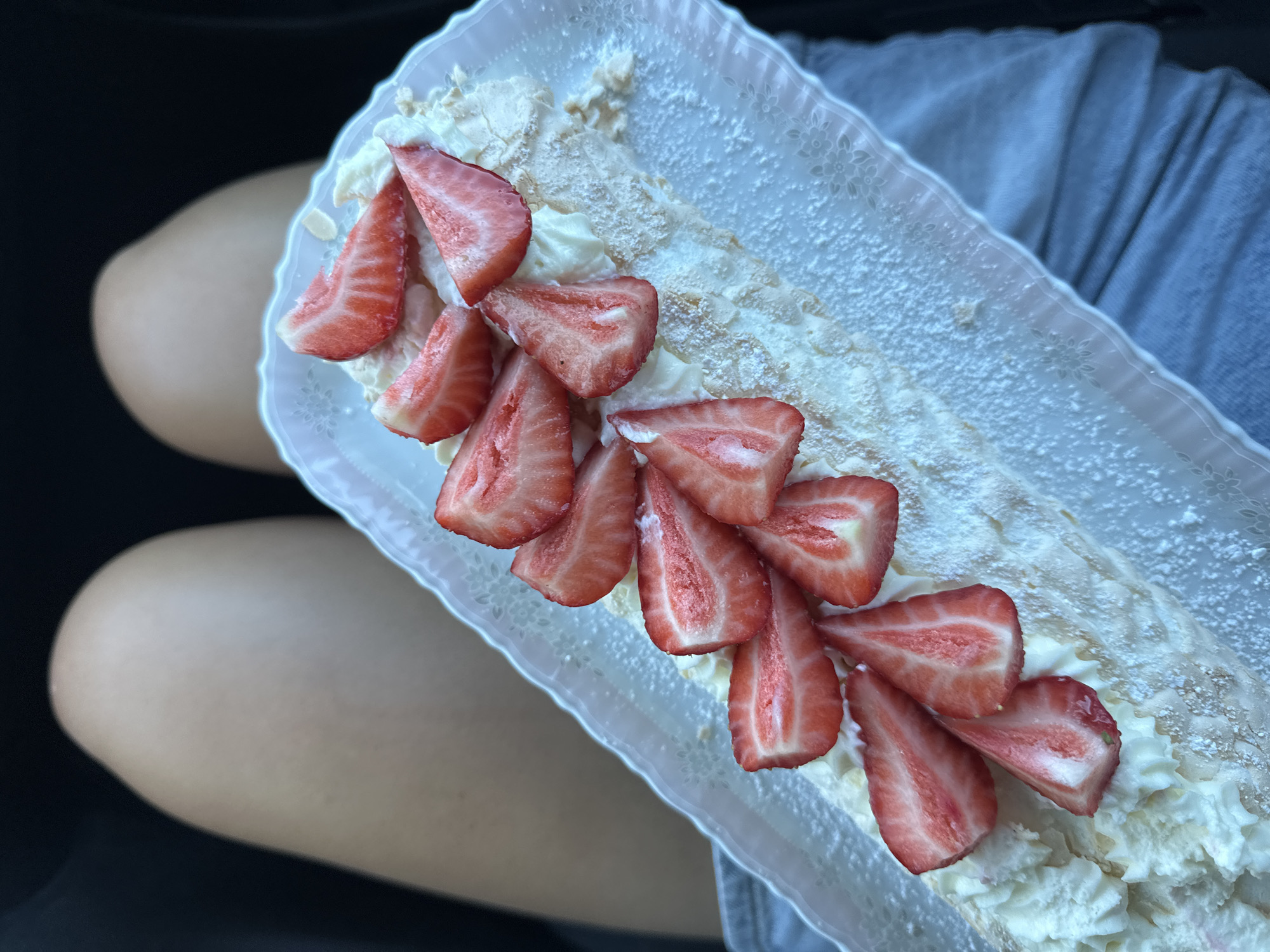 The image shows a dessert, specifically a meringue roll topped with sliced strawberries and powdered sugar, resting on a person's lap. The person is wearing a gray shirt. The dessert is in a clear, disposable rectangular tray. The background appears to be inside a vehicle. There is no indication that the image was taken in Kutenholz, Germany.