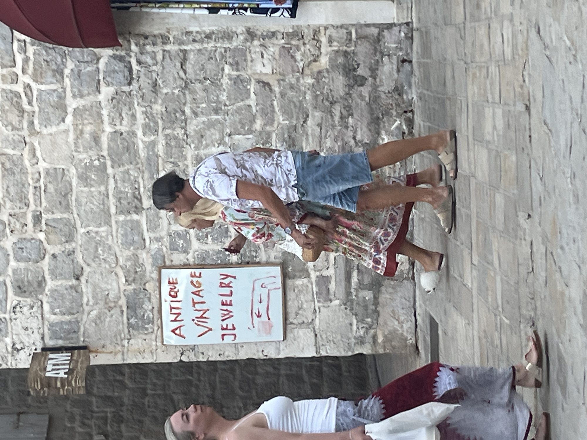 The image depicts a street scene in the old town of Budva, Montenegro. In the foreground, a woman with blonde hair is walking from left to right, wearing a white tank top and a long skirt with red and white patterns. She is carrying a white bag in her left hand. To her right, a couple is walking in the same direction. The man has dark hair and is wearing a patterned short-sleeved shirt and blue denim shorts. The woman next to him has blonde hair and is wearing a colorful floral dress. The couple appears to be walking close together. Behind them, a white sign with red lettering reads "ANTIQUE VINTAGE JEWELRY" and has a simple arrow diagram. Above the sign, a wooden sign with "ATM" is visible. The buildings in the background are constructed of grey stone blocks. The street is paved with square stones. The overall scene suggests a warm, possibly summer day in a historic European town.