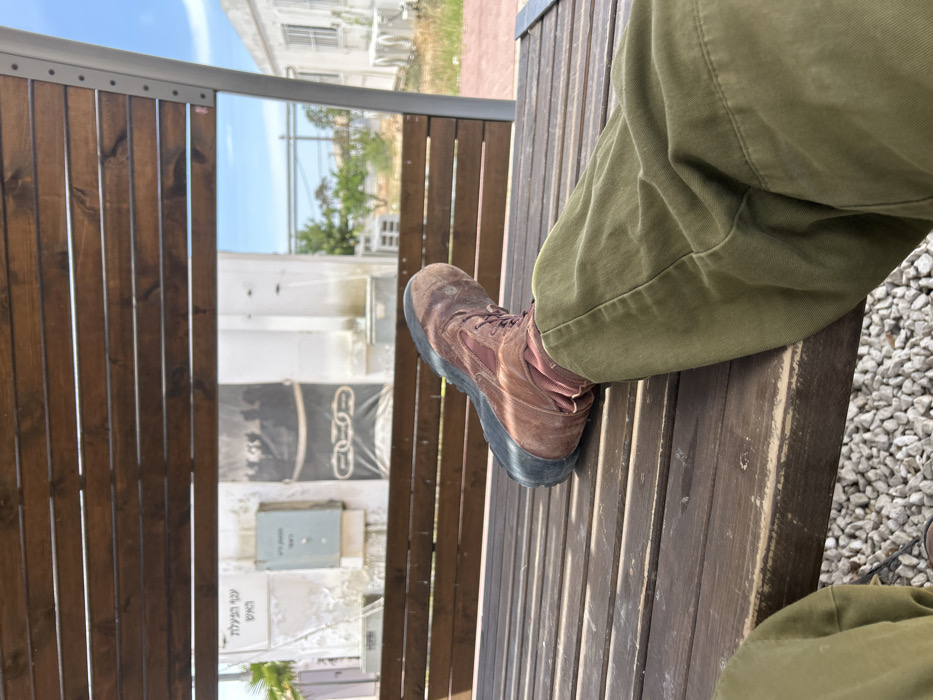 A brown leather boot rests on a wooden bench with the leg of the wearer, clad in olive green pants, resting on the bench. The bench is under a wooden pergola, and a white building is visible in the background. The setting appears to be a park or outdoor public space. The coordinates 31.173, 34.559 correspond to a location in Tze'elim, Israel. The wooden structure has Hebrew writing on it.
