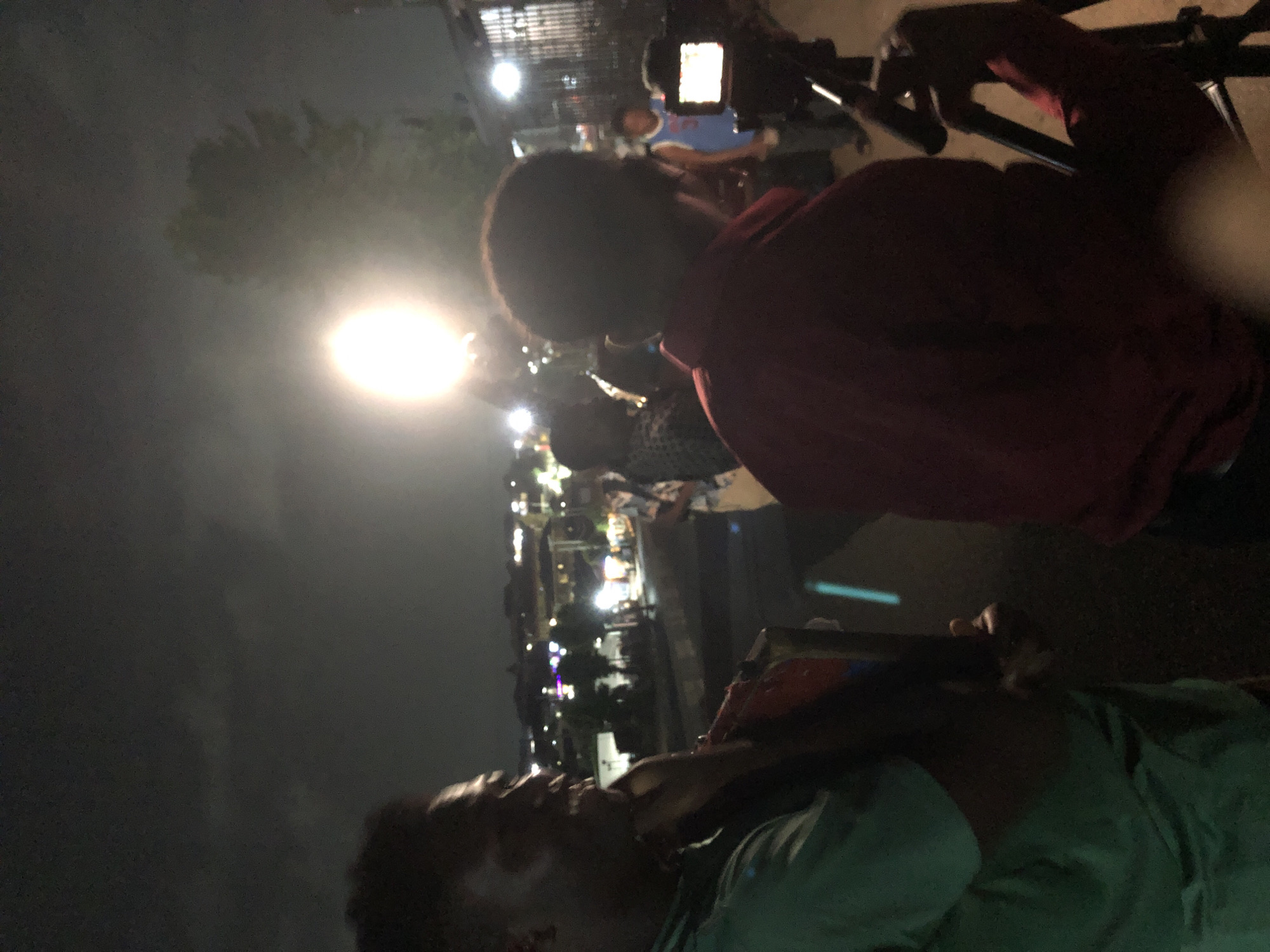 This media file captures a nighttime outdoor scene, likely an event or public gathering, characterized by artificial lighting and media activity. The image is rotated 90 degrees clockwise, with the horizon on the left and right.
In the foreground, from a slightly low angle, a person wearing a light green or teal shirt is visible, appearing to lean back or look upwards. Their face is partially illuminated, showing some facial features.
Immediately behind and slightly above them, another person, wearing a dark red or maroon long-sleeved top, is positioned, seen from behind and to the side. This individual is actively operating a camera mounted on a tripod, which is facing away from the viewer. The screen of the camera is lit, suggesting it is powered on and in use. The hands of the person in red are gripping the camera or tripod handles.
The midground and background are dark but illuminated by several bright artificial light sources. A prominent, intensely bright, circular light source is visible on the left side of the frame (when correctly oriented), casting a strong glow. Other scattered lights in the distance suggest a street, market, or an area with multiple structures and activity. Faint outlines of trees or foliage are also discernible against the dark sky in the upper left. Several other individuals are present in the background, though their features are indistinct due to the low light and distance, contributing to the sense of a crowd or gathering.
The overall impression is that of a public event being filmed or documented after dark. No legible text is visible.