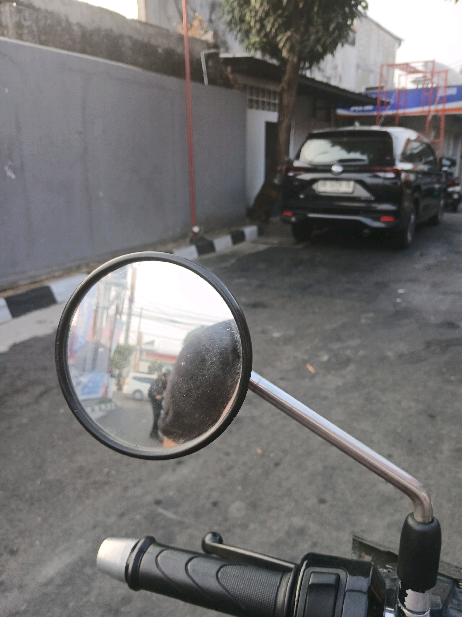 The image captures a street scene in Depok, Indonesia, as viewed from a motorcycle. In the foreground, the motorcycle's handlebar and a portion of the brake lever are visible. A circular rearview mirror is prominently featured, reflecting a blurred image of the surroundings.
The reflection in the mirror shows a street with a black car parked or moving, a portion of a building with a blue and orange sign, a tree, and overhead wires. A person, likely the rider, is partially visible as a reflection in the mirror, with dark hair. A second person, wearing dark clothing and a helmet, is also reflected, standing near the black car.
The background of the image displays a grey wall on the left, a tree, a white building, and a red structure that appears to be part of a rack or scaffolding. The street surface is paved and appears somewhat worn. The lighting suggests it is daytime, with sunlight casting shadows. The presence of overhead wires and the architectural style of the buildings are characteristic of urban environments in Indonesia.