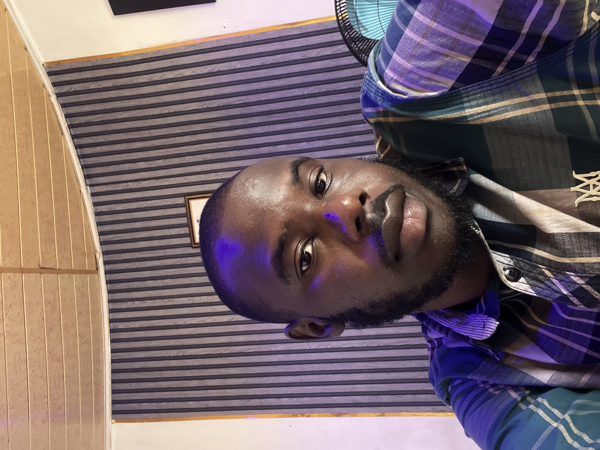 A man is seen in a close-up selfie, looking directly at the camera. He has dark skin, a shaved head, and a beard. He is wearing a plaid shirt with various shades of blue, green, and brown. The background consists of a striped wall with a wooden ceiling above it. To the right, a blue fan is partially visible. The lighting in the scene is dramatic, with purple and pink hues cast on the man and the background, suggesting an artificial light source. The image appears to be taken indoors, possibly in a home or office setting. The location of Jos, Nigeria, is not explicitly indicated in the image itself and cannot be determined from the visual information provided.