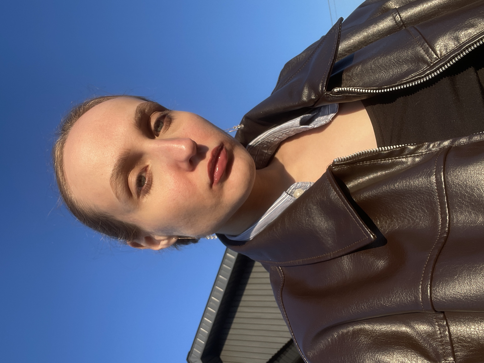 A close-up portrait captures a young person, likely female, looking directly at the camera. She has fair skin, her hair is pulled back, and she has soft, natural-looking eyebrows and glossy, reddish-brown lips. She wears a brown, textured, leather-like jacket with visible zippers and seams, layered over a black top and a striped collar of a button-down shirt. A delicate, chain-like earring is visible on her left ear.
The photograph appears to be a selfie or a low-angle shot, taken outdoors on a bright, sunny day. The dominant background is a clear, deep blue sky, indicating fair weather. Direct sunlight casts strong highlights on her skin and the shiny material of her jacket, suggesting a low sun angle, possibly morning or late afternoon. In the lower right corner, a portion of a light-colored building with vertical siding or roofing is visible, contrasting with the expansive sky.
The subject's expression is neutral and direct, creating a contemplative or posed atmosphere. There is no visible text within the image. The setting is in Подшивалово, Russia.