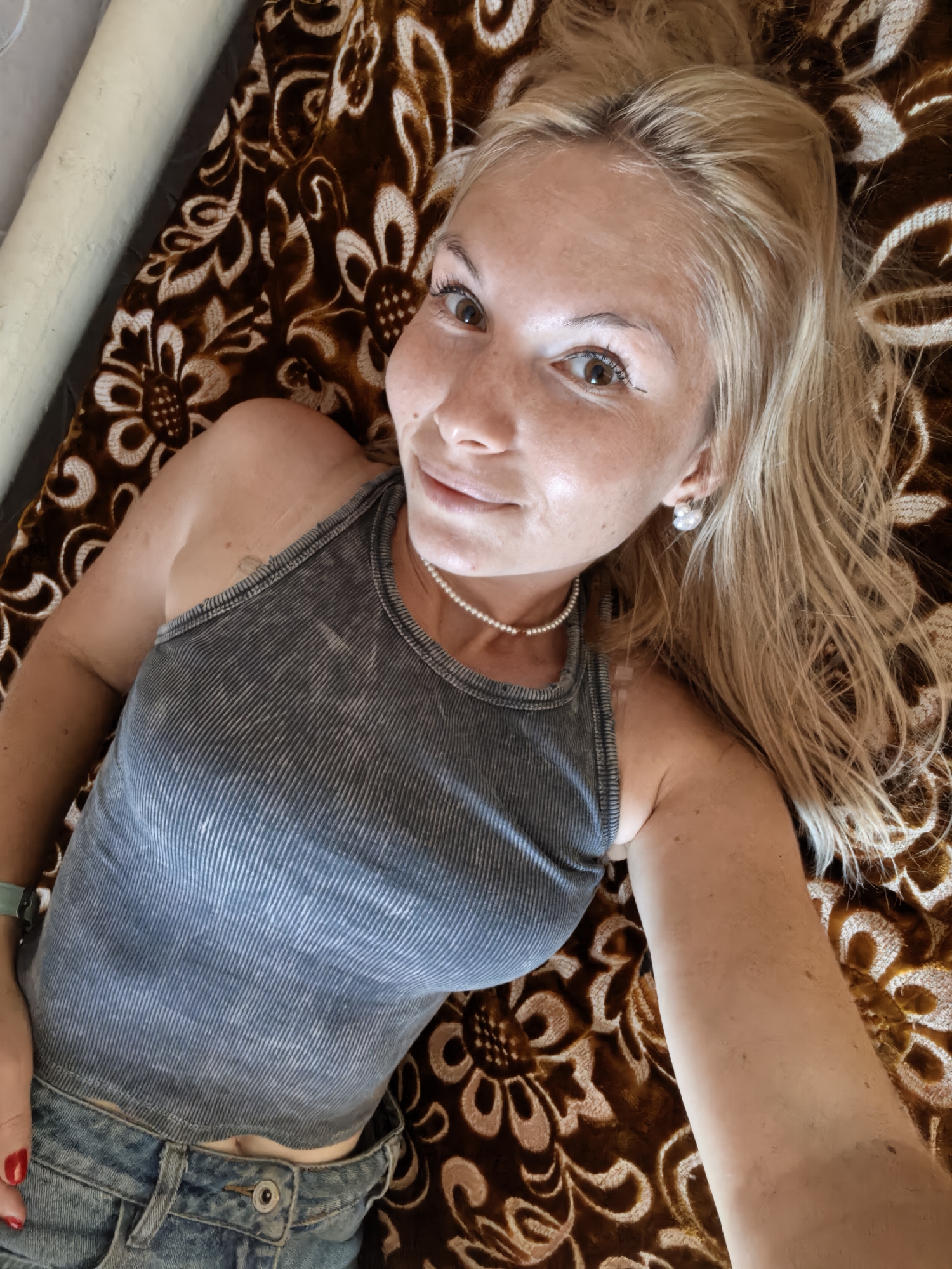 A woman with blonde hair and freckles is lying on a brown and white patterned blanket. She is wearing a grey tank top and blue jeans. She is looking at the camera with a slight smile. The image appears to be a selfie. There is no information in the image that indicates the location in the city of Sterlitamak, Russia.