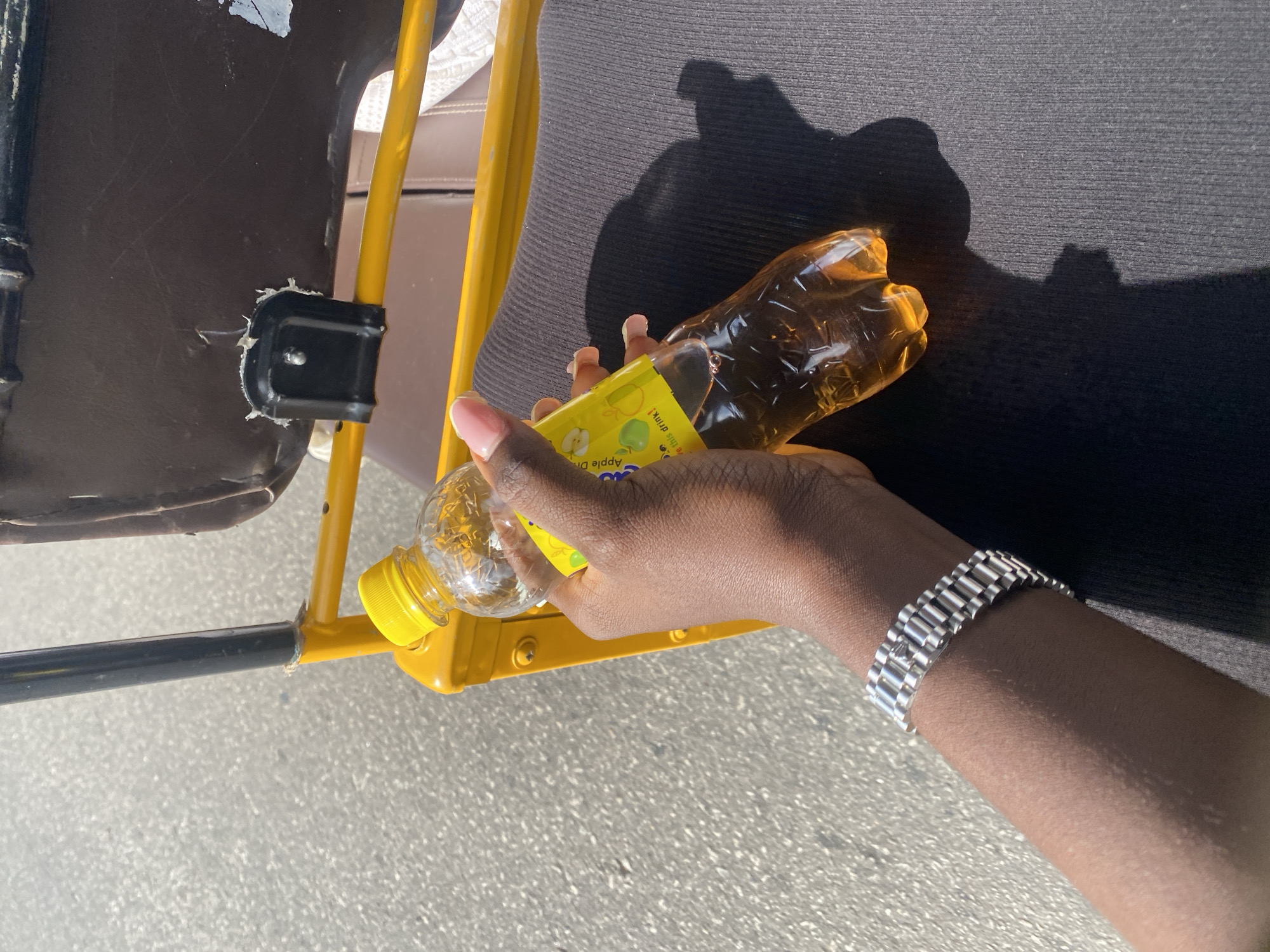 A close-up shot captures a dark-skinned hand holding a clear plastic bottle of amber-colored liquid. The bottle has a yellow cap and a label featuring green apples and the words "Apple Drink." The hand is adorned with a silver-toned watch. The person is seated on what appears to be a yellow and brown vehicle seat, with a dark gray ribbed fabric visible beneath. The background is an asphalt surface, suggesting an outdoor urban setting. The scene is lit by direct sunlight, casting a distinct shadow of the bottle and hand. The image was taken in Jos, Nigeria.