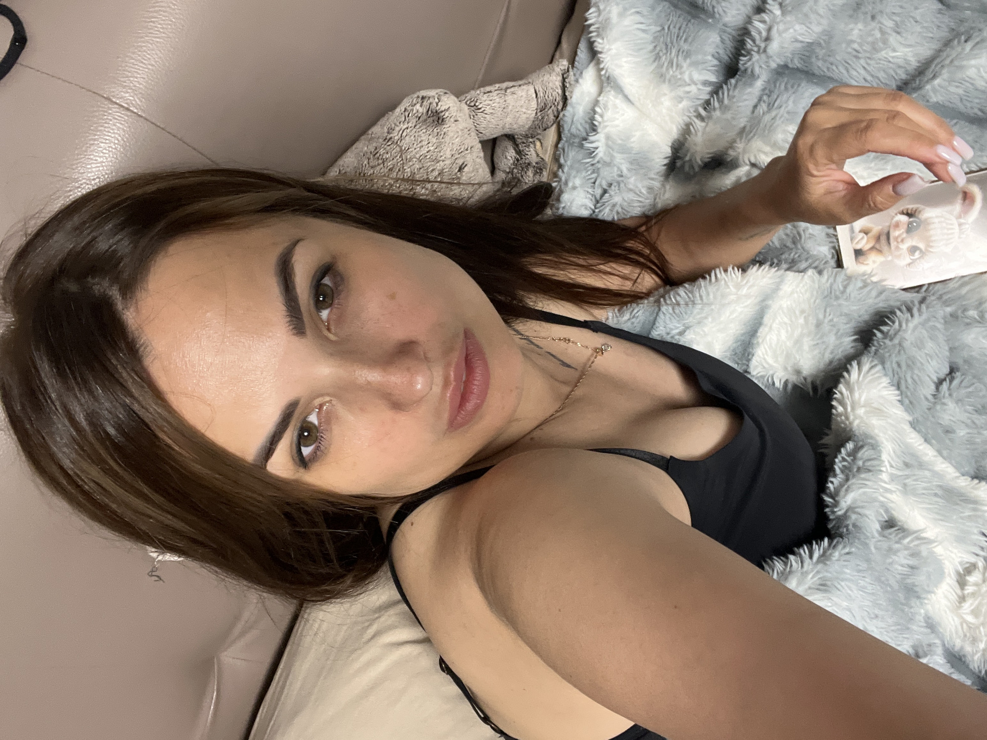 A young adult female with medium-length brown hair and brown eyes is lying down, propped up by pillows on a light-colored sofa. She is wearing a black bra and a necklace with a pendant. Her skin is fair with freckles. She is holding a phone in her right hand and a gray and white fuzzy blanket is draped over her. The background is a soft, out-of-focus beige wall and sofa. The lighting appears to be warm and artificial, possibly from an indoor light source.