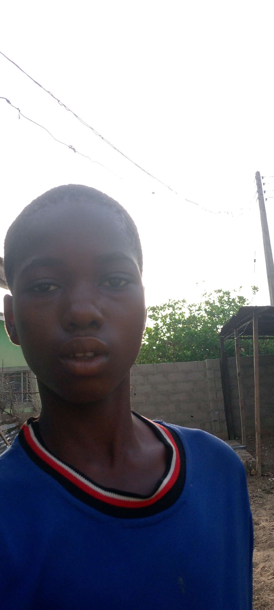 A close-up shot of a young Black male in Adankolo, Nigeria, wearing a blue shirt with a black, red, and white striped collar. A cinder block wall and green foliage are visible in the background. A wooden structure with a roof stands to the right. A utility pole and wires are also visible in the upper background. The lighting indicates daytime.