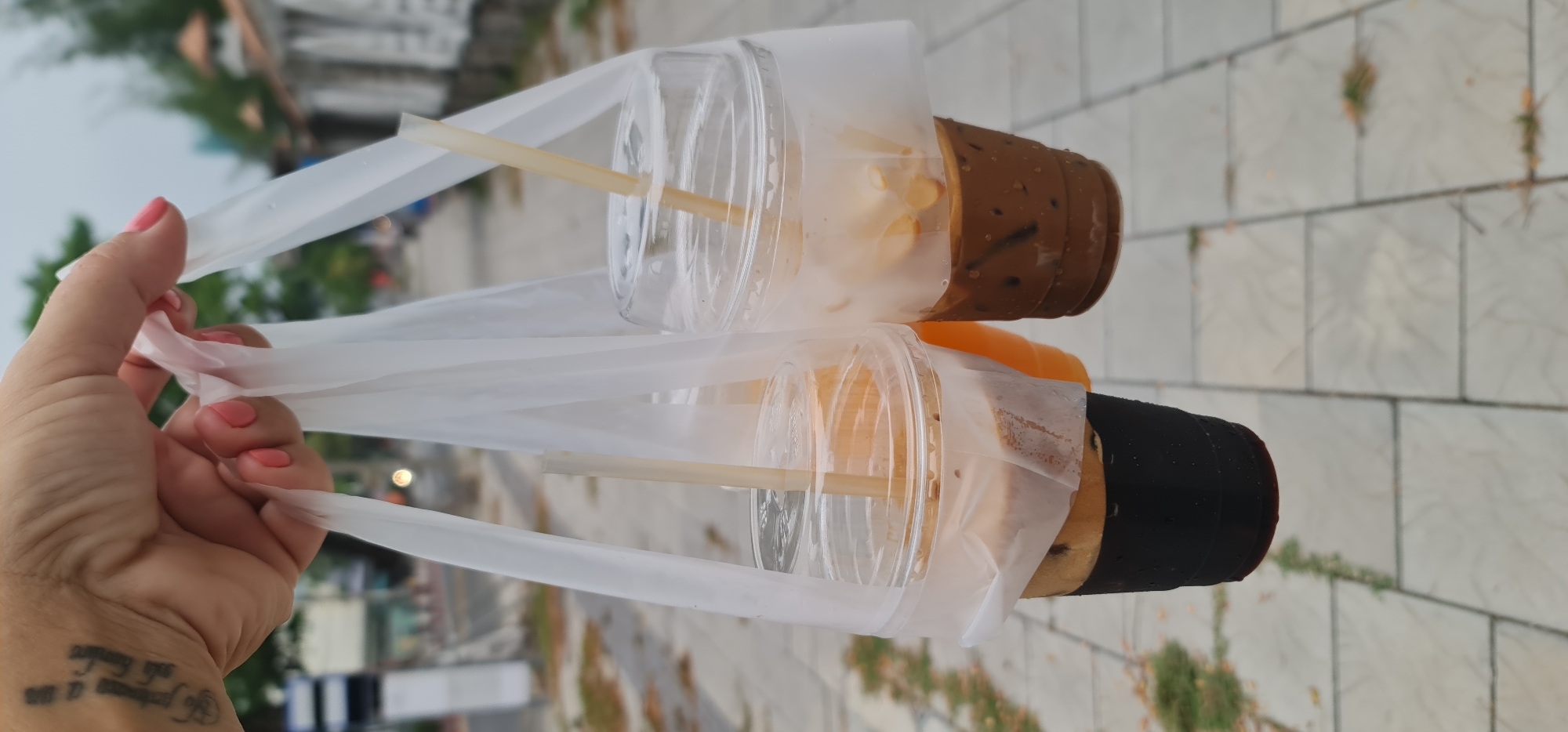 A person holds two plastic bags, each containing a beverage, in front of a tiled walkway. The upper drink is a light brown coffee with ice in a clear plastic cup with a straw. The lower drink appears to be orange juice over a dark base, also in a clear plastic cup with a straw. The person holding the drinks has a visible tattoo on their wrist and pink nail polish. The background shows a city street with trees and buildings. The image location is implied to be Đà Nẵng, Vietnam.