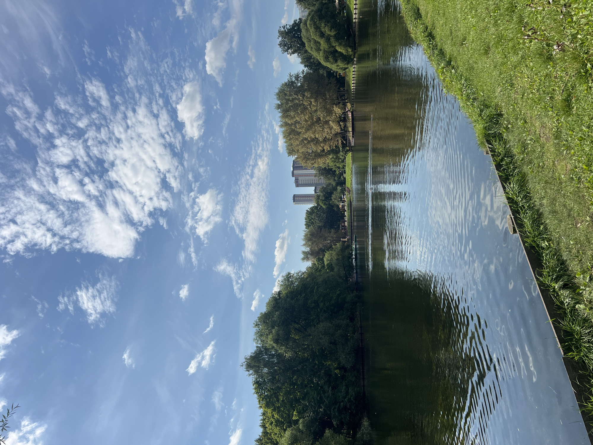 The image captures a serene view of a park in Moscow, Russia, on a bright, partly cloudy day. The scene is dominated by a tranquil body of water, reflecting the blue sky and scattered white clouds. Lush green trees line the far bank of the lake, creating a vibrant natural backdrop. In the distance, several tall, modern buildings rise above the tree line, indicating an urban setting within the park. The foreground features a neatly kept grassy embankment sloping down to the water's edge. A wooden border runs along the edge of the grass, separating it from the lake. Several small boats, possibly paddle boats, are visible on the water near the opposite shore. The overall atmosphere is one of peaceful recreation within an urban park environment.