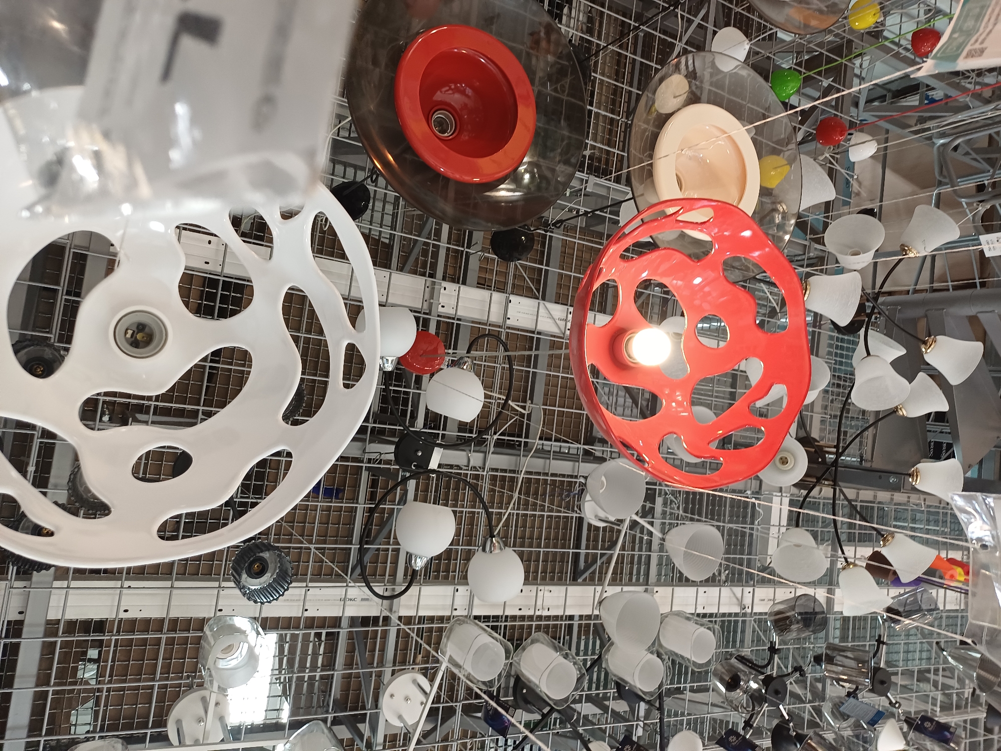 The image displays a low-angle shot looking up at a ceiling filled with a variety of light fixtures, suspended from a metal grid. The lighting is varied, with some fixtures appearing to be illuminated, while others are not. The scene appears to be an indoor retail environment, likely a home improvement or lighting store, given the abundance and variety of display items.
Specific objects visible include:
- Several white pendant lights with rounded shades.
- A large, white, circular fixture with an intricate, cut-out design.
- A red, spherical fixture with an opening that reveals an illuminated bulb.
- A dark, almost black, circular fixture with a prominent red interior, possibly a spotlight or downlight.
- Various smaller glass or plastic light shades in white and clear.
- Some bare bulbs are visible.
- Metal components and wiring are present, indicating the display and operational nature of the fixtures.
- The ceiling grid is a prominent feature, acting as the mounting system for the lights.
The overall impression is one of a brightly lit retail space dedicated to lighting products. There are no people or animals visible in the image. The location of Boryspil, Ukraine, is not deducible from the visual information provided in the image itself.