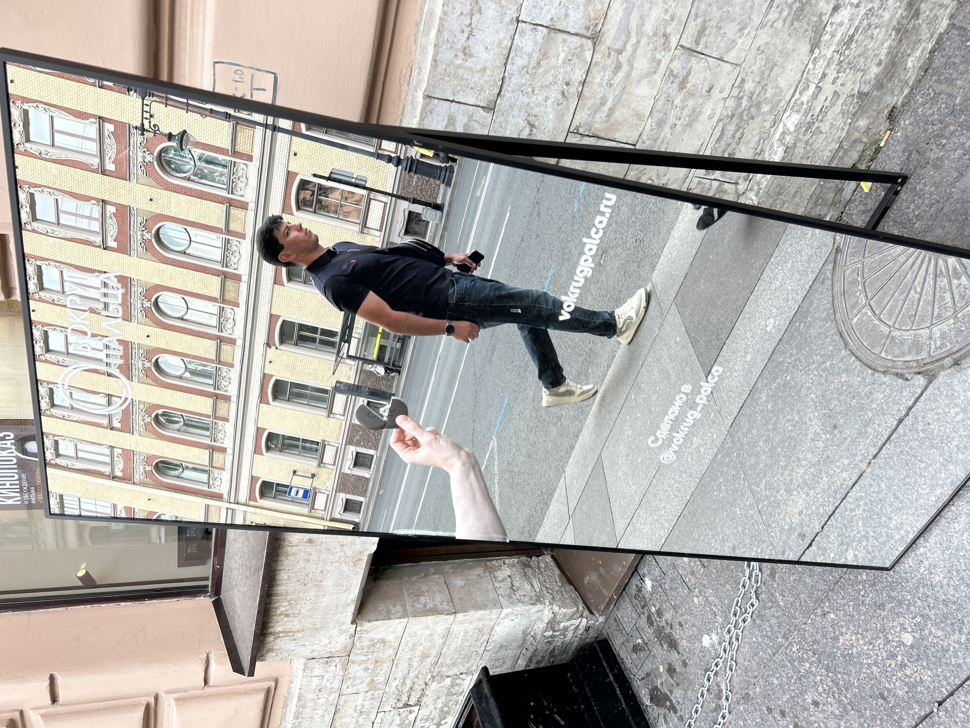 A tall, rectangular mirror signboard, framed in thin black metal, stands on a light gray concrete sidewalk in an urban setting. The mirror is angled, reflecting a street scene and a pedestrian.
In the reflection:
A male pedestrian, with short dark hair, wearing a dark blue polo shirt, dark blue jeans, and light-colored sneakers, walks from the viewer's right to left across a paved street. He carries a black shoulder bag and holds a black smartphone in his right hand, with a dark smartwatch on his left wrist. Behind him, a multi-story building with a distinctive yellow and reddish-brown brick facade features numerous arched windows with white decorative framing. A dark, ornate streetlamp is positioned in front of this building, and a blue square bus stop sign with a white bus icon is visible at street level. White Cyrillic text, reading "Сделано в @vokrug_palca.ru", is reflected on the sidewalk in the lower foreground. A disembodied light-skinned arm extends from the left edge of the reflection, holding a dark, hinged, and irregularly shaped object open, revealing a lighter interior.
Directly visible elements surrounding the mirror:
To the left of the mirror, a building wall is visible. Its upper section is pale peach-colored with decorative molding, featuring a dark window frame with an interior spotlight and a dark brown metal ledge below. The lower part of this wall is constructed from textured, light-colored stone blocks, with an opening at ground level. A black metal chain lies coiled on the concrete sidewalk in the lower left foreground. A circular metal manhole cover with a radial spoke design is set into the concrete sidewalk in the lower right. A discarded cigarette butt lies on the ground near the base of the mirror's black support leg. A partial black structure, possibly a waste bin, is situated at the far lower left next to the stone wall. The scene is illuminated by daylight.