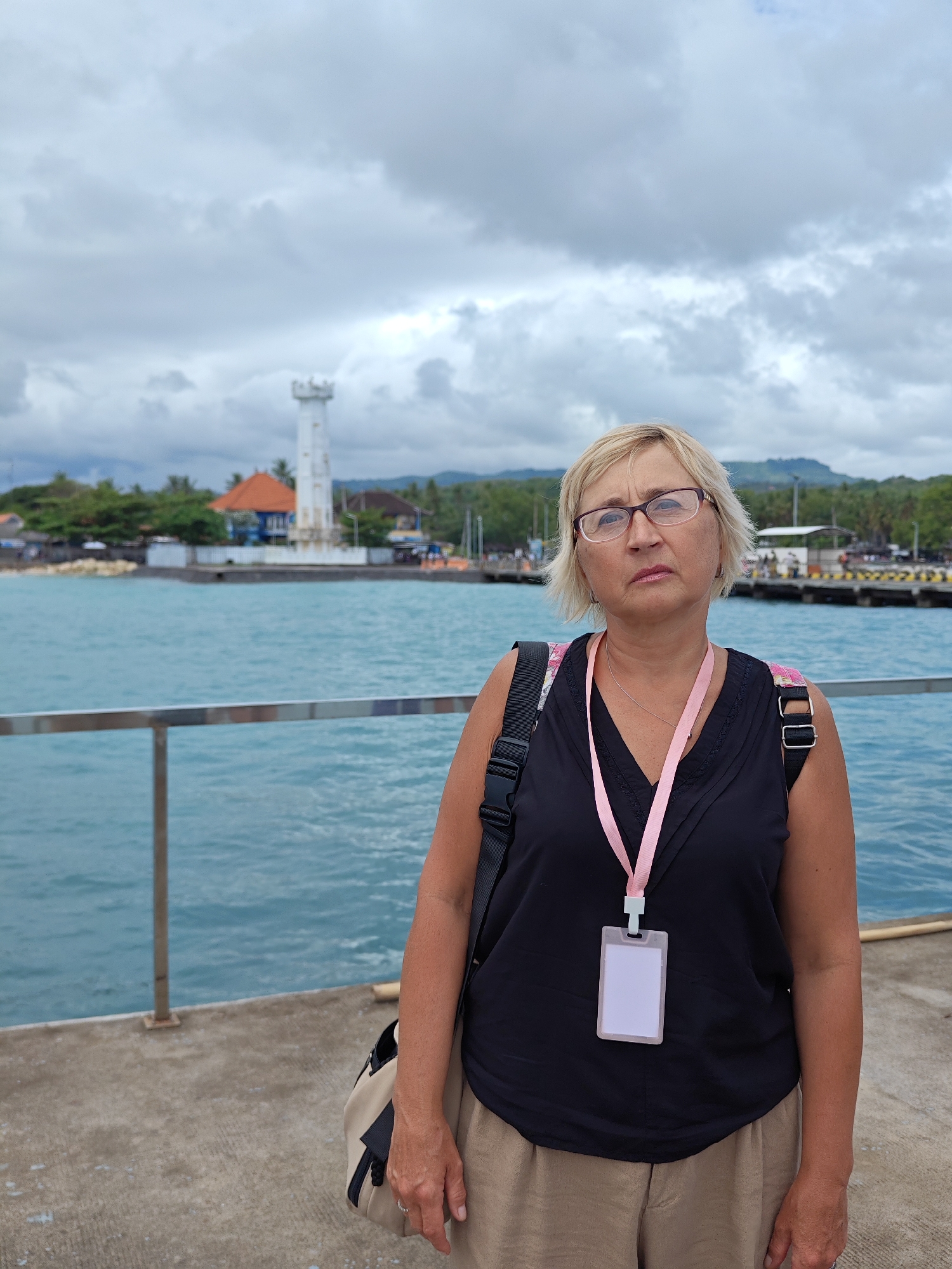 A medium shot depicts a middle-aged woman with blonde hair and glasses, standing on a concrete pier. She wears a black sleeveless top, beige pants, and carries a bag on her shoulder. A lanyard with a blank, white ID card hangs around her neck. Behind her, the turquoise ocean stretches out, with a clear view of a white lighthouse and buildings characteristic of the coastal area. The background features cloudy skies and distant hills. The scene is set in Kampung Toyapakeh, Indonesia, evidenced by the architectural style and coastal environment.