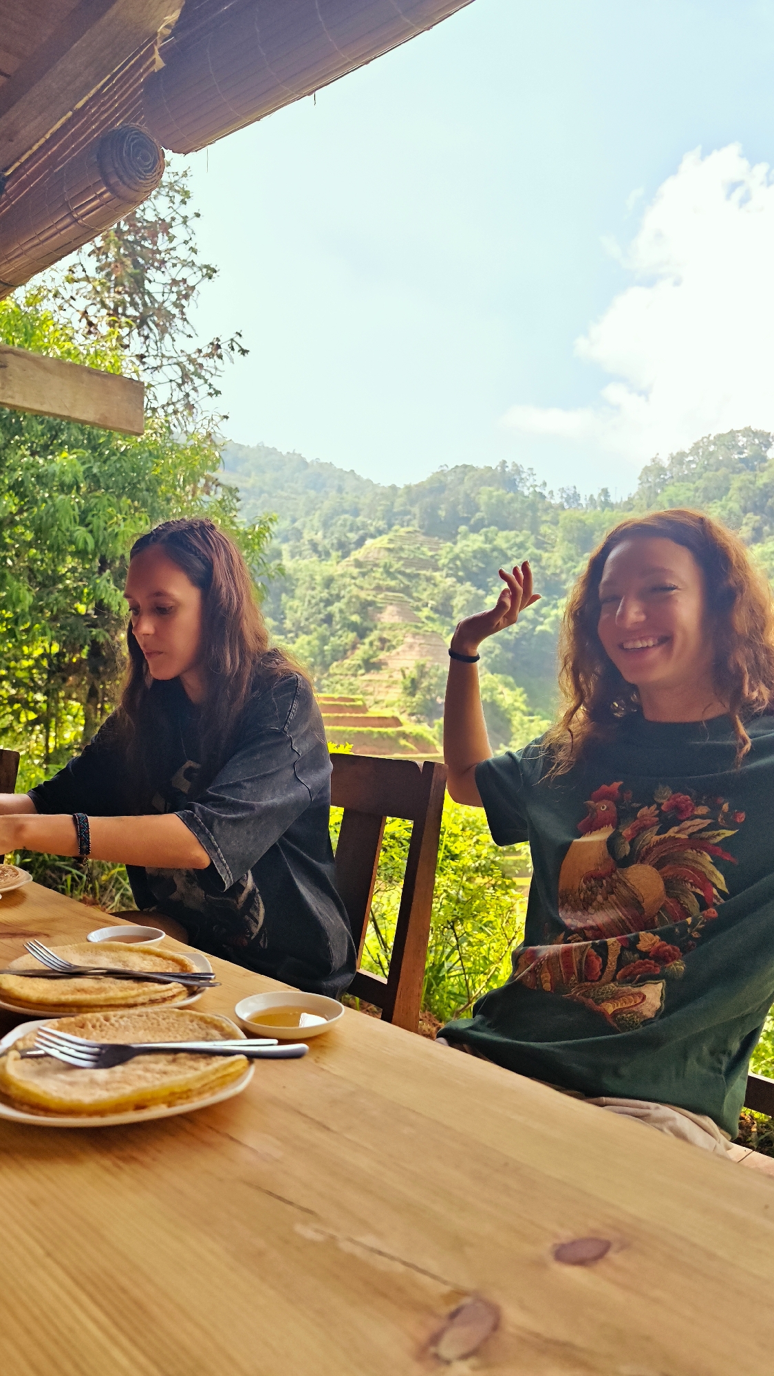 Two Caucasian women are seated at a wooden table on a veranda overlooking a rice paddy field in Hoàng Su Phì Commune, Vietnam. The woman on the left has dark hair and is wearing a black shirt. She is looking down at her plate of pancakes. The woman on the right has red hair and is wearing a green shirt with a rooster design. She is smiling and has her right hand raised. There are plates of pancakes with forks and small bowls of honey on the table. The background shows lush green hills and terraced rice fields. The sky is blue with some clouds. A bamboo shade is visible at the top of the frame.