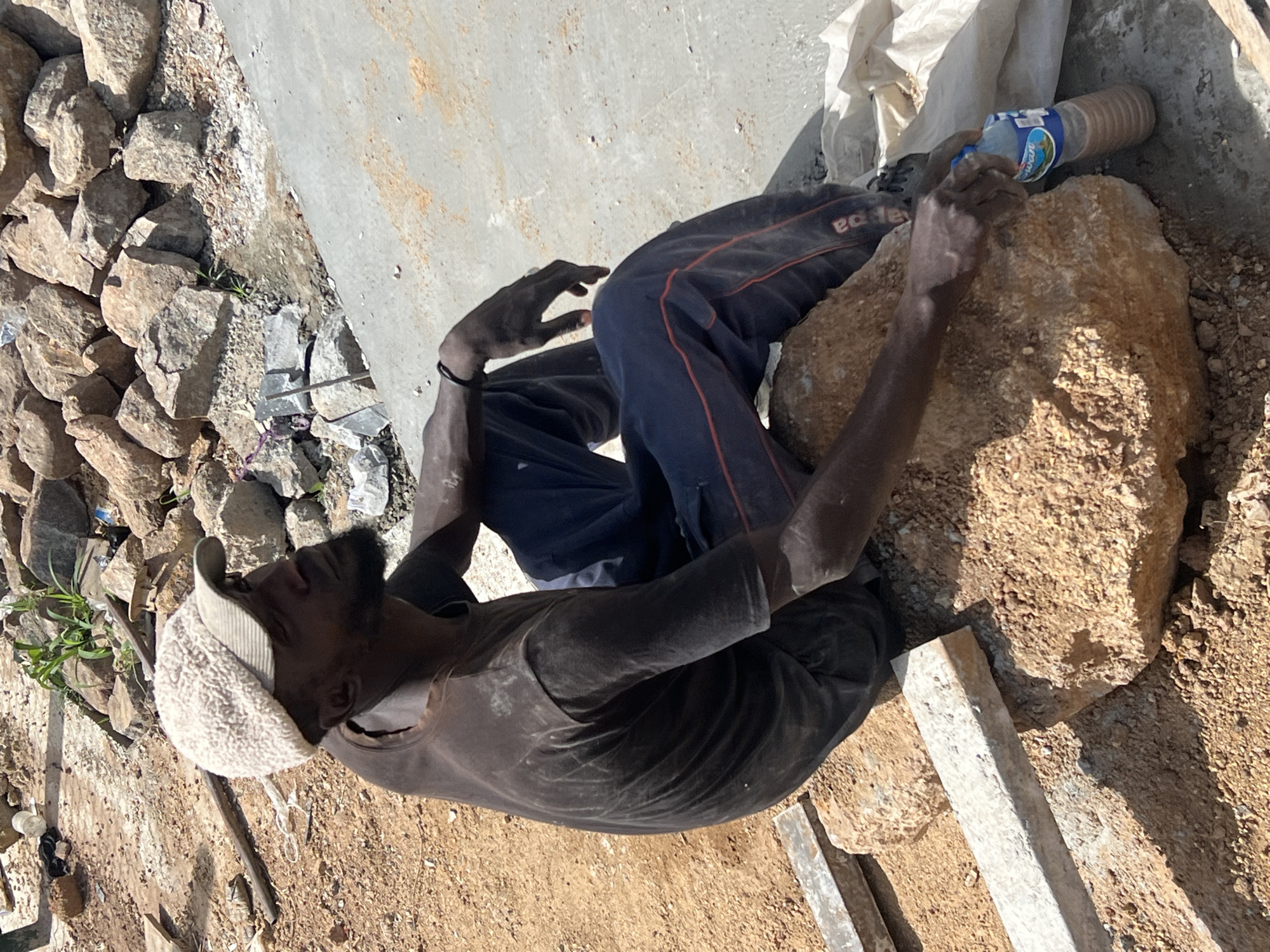 A man in Barakin Lerawi, Nigeria, rests outdoors on a large stone. He wears a cap, dark shirt, and dark pants with a red stripe. He holds a plastic water bottle. Rocks surround a concrete slab. Metal bars lie nearby on the dirt ground.