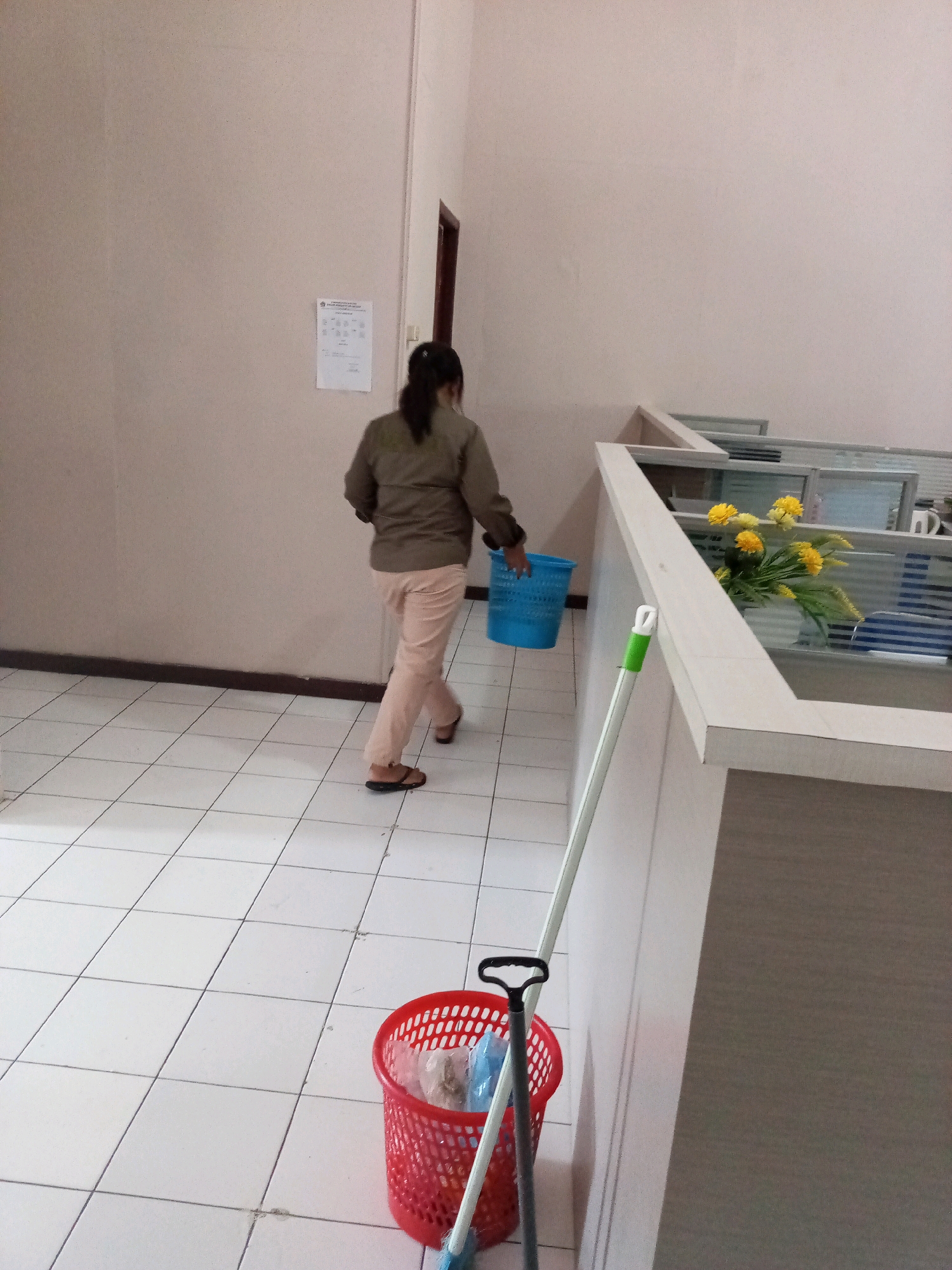 The image depicts an indoor scene, likely an office environment, with white tiled flooring and pale pink walls. A woman with dark hair pulled back is walking away from the camera, wearing a green jacket and light pink pants, with brown sandals. She is carrying a blue plastic basket. In the foreground, a red plastic basket filled with various items sits on the floor next to a broom with a green handle. To the right, a low partition wall, likely part of office cubicles, is visible. Yellow flowers in a vase are placed on top of this partition. There is a door visible on the far wall. A notice is pinned to the wall on the left. The scene appears to be located within the city of Bitung, Indonesia, as indicated by the request.