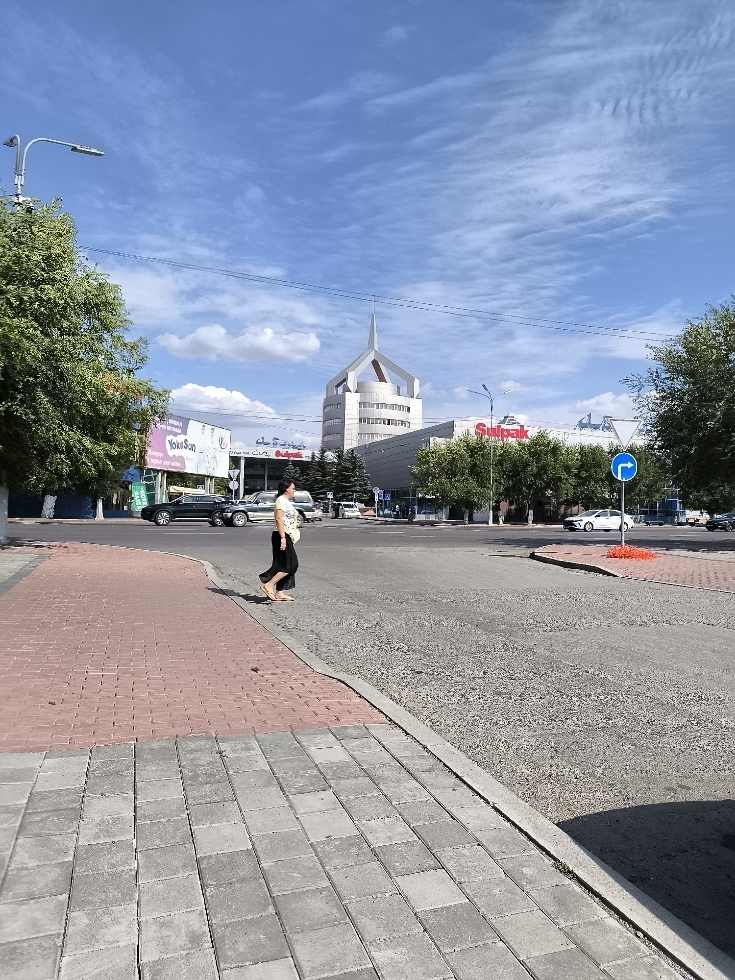 A woman with dark hair, wearing a light-colored patterned shirt and a black skirt, walks across a street at a pedestrian crossing. She is positioned in the middle of the street, moving from left to right in the frame. A white car is visible in the background on the right side of the road, and another car is stopped at the crossing to her left. The street is paved, and the pedestrian crossing is marked with white lines. The sidewalk on the left side of the street is made of light gray and red bricks. In the background, there is a modern building with a distinctive pointed roof, signage that reads "Sulpak" in red letters, and other commercial buildings. Trees are planted along the sidewalks and in front of some of the buildings. The sky is blue with scattered white clouds. A street lamp is visible on the left side of the image, near the edge of the frame. The scene appears to be a daytime street view in a city.