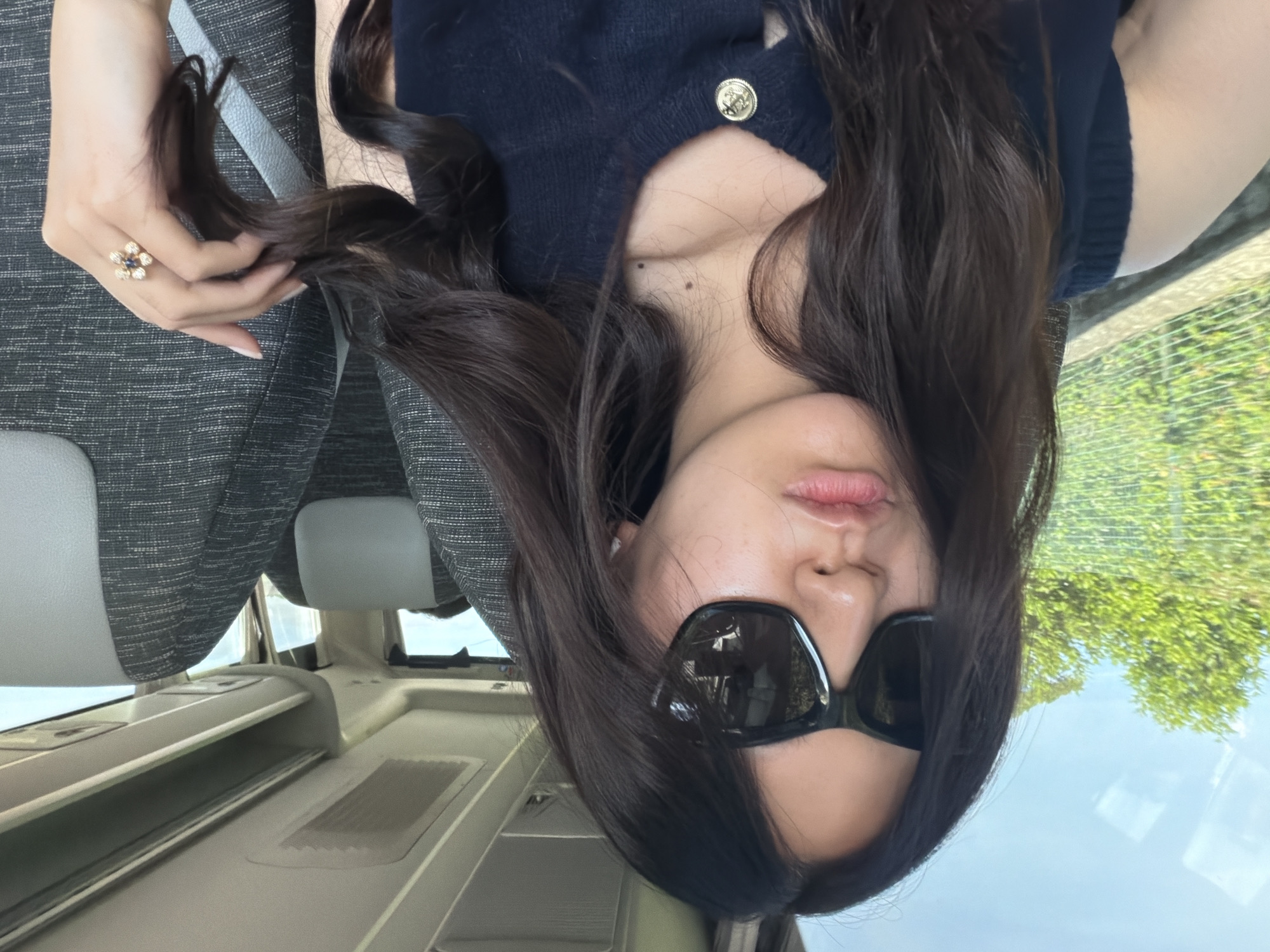 A woman with long, dark hair, wearing dark sunglasses and a dark top with a gold button, is posing inside a vehicle. She is holding a strand of her hair and wearing a ring on her finger. Outside the vehicle, green trees can be seen through the window. The interior of the vehicle appears to be white and gray. There is no definitive information to confirm that the location is in Saint-Tropez, France.