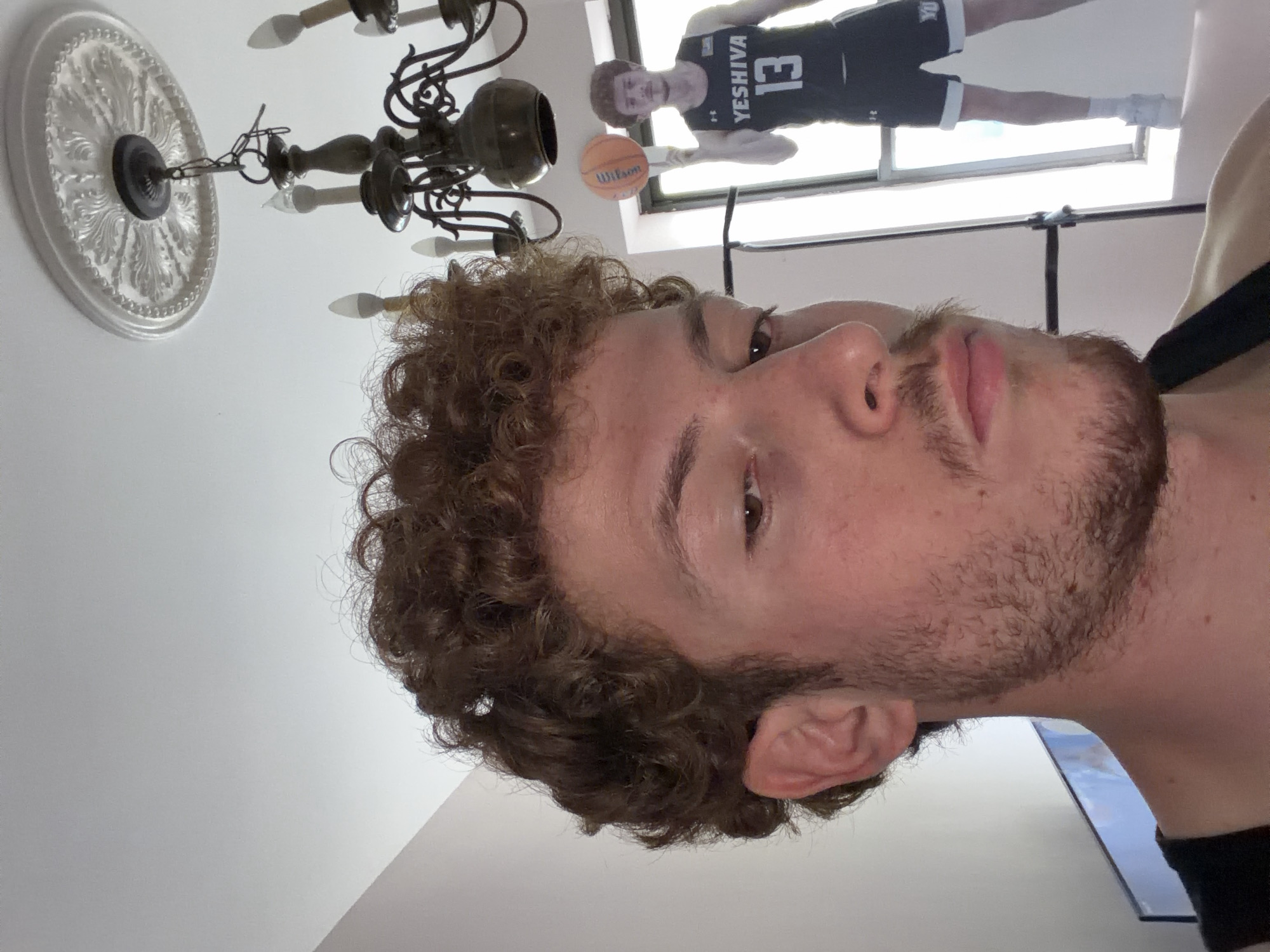 A close-up portrait, oriented sideways, features a young man with curly light brown hair, a short beard, and brown eyes, looking towards the camera with a neutral expression. The image captures him from a slightly low angle, showing his face and the side of his head.
In the background, a reflection of a person, likely the same individual, is visible in a window or large mirror. This reflected figure is standing, wearing a black basketball jersey with "YESHIVA" printed across the chest and the number "13" below it. White shorts and white sneakers complete his attire. He is holding an orange "Wilson" basketball in his left hand, positioned as if in a basketball stance. The bright light surrounding the reflected figure suggests daylight streaming in from outdoors.
Above the main subject, a decorative, ornate white ceiling medallion is visible, from which a dark, multi-armed chandelier hangs down. The chandelier has several light fixtures resembling candles. The ceiling itself is white. The scene appears to be an indoor setting, possibly a residence, with the light from the background indicating it is daytime. The "YESHIVA" text on the jersey suggests a connection to Yeshiva University, aligning with the given location context of New York, United States of America.