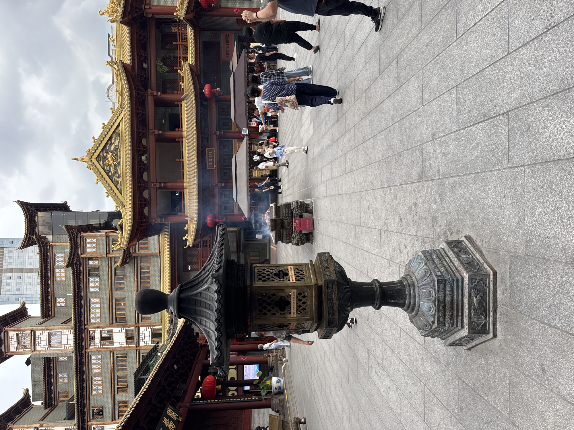 The image captures a scene in Yuexiu District, China, showcasing a traditional building complex on a stone-tiled plaza. The foreground features an ornate metal lantern or incense burner with intricate detailing. A line of people stands in the background, likely tourists or worshippers. Smoke rises from burning incense or offerings in designated areas. The buildings exhibit traditional Chinese architectural styles with tiered roofs, golden accents, and red lanterns. Modern buildings are visible in the background, juxtaposing the old and new. The sky is overcast, suggesting a cloudy day.