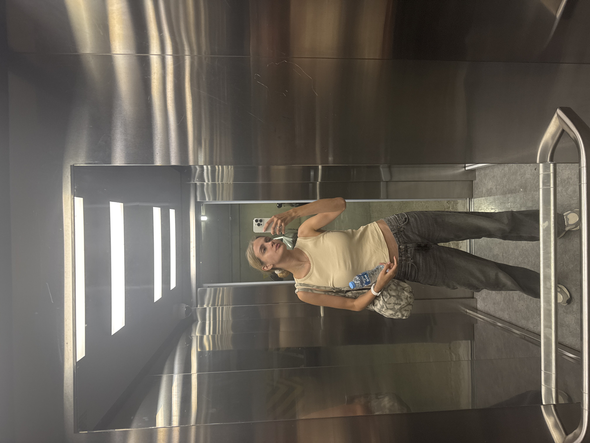 A young woman is reflected in a large mirror inside an elevator. She is facing the mirror and holding a cell phone in her right hand, presumably taking a selfie. She is wearing a tan tank top, dark-colored jeans, and sneakers. She also has a bag on her left shoulder and is holding a bottle of water in her left hand. The elevator's interior consists of stainless steel panels and bright vertical light fixtures. The location is unknown.