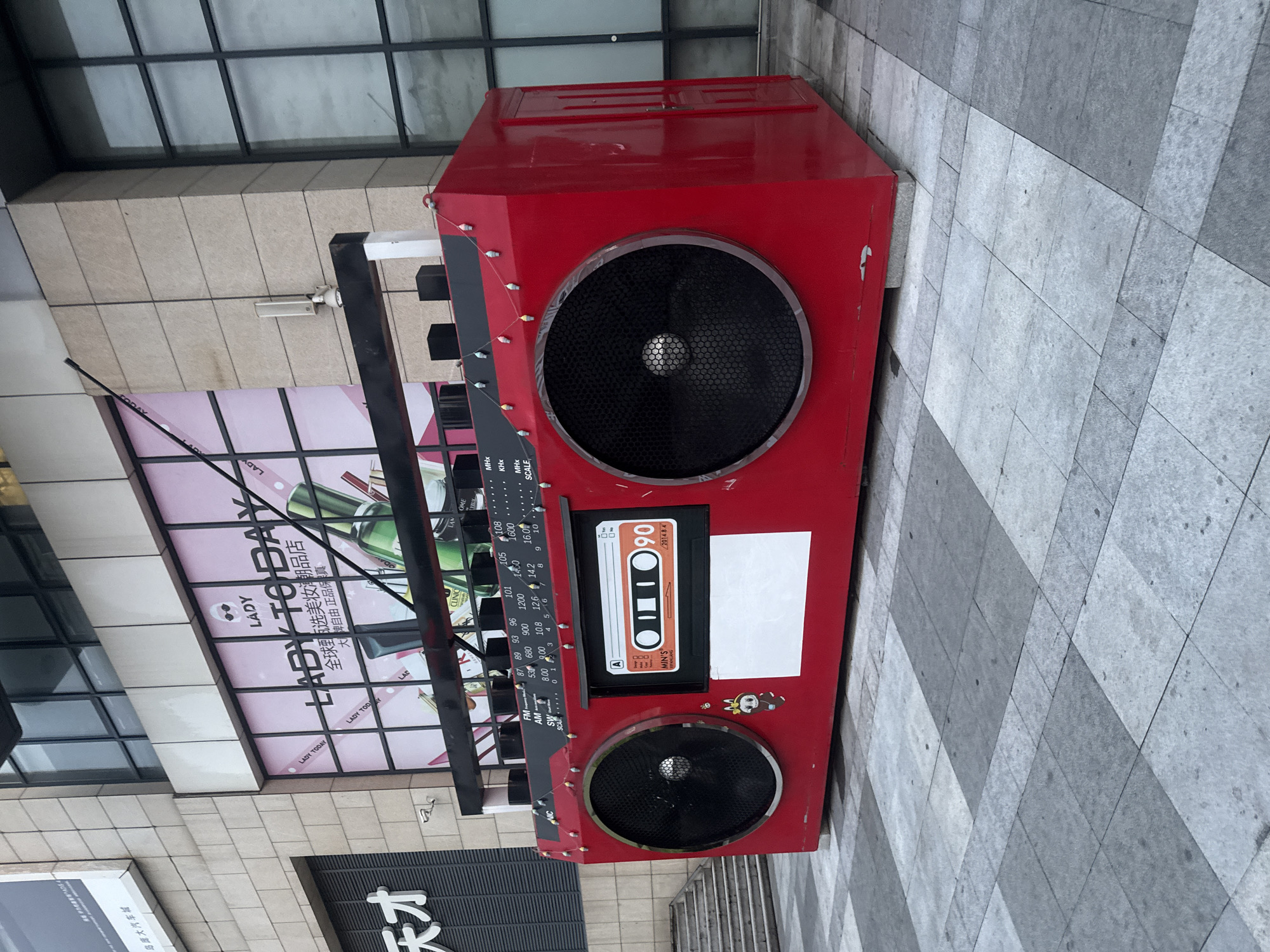 This is a medium shot of a large, decorative boombox sculpture displayed outdoors in front of a building. The boombox is bright red and features two large speakers, a tape deck with a cassette displaying "90", and various control knobs and dials. It is adorned with small, colorful lights strung along the top. In the background, a building facade is visible, featuring large windows and signage that reads "LADY TO DAY" in English and Chinese characters. The ground in the foreground is paved with large, square grey tiles arranged in a checkerboard pattern. The overall scene appears to be a public space in a city. Specifically, this sculpture is located in the city of Qinhuangdao, China, outside a Lady to Day store.