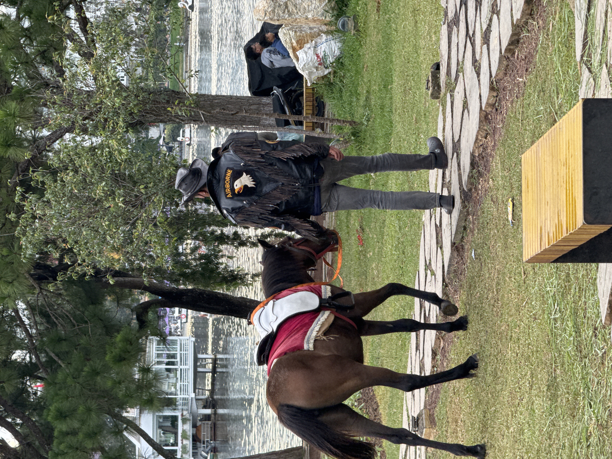 A man in a cowboy hat and a black leather jacket with fringe and an "AIRBORNE" patch walks on a paved path beside a lake in Da Lat, Vietnam. He is leading a brown horse with a red saddle. In the background, there is a white building with large windows and several people sitting near the water. Trees and greenery surround the scene.