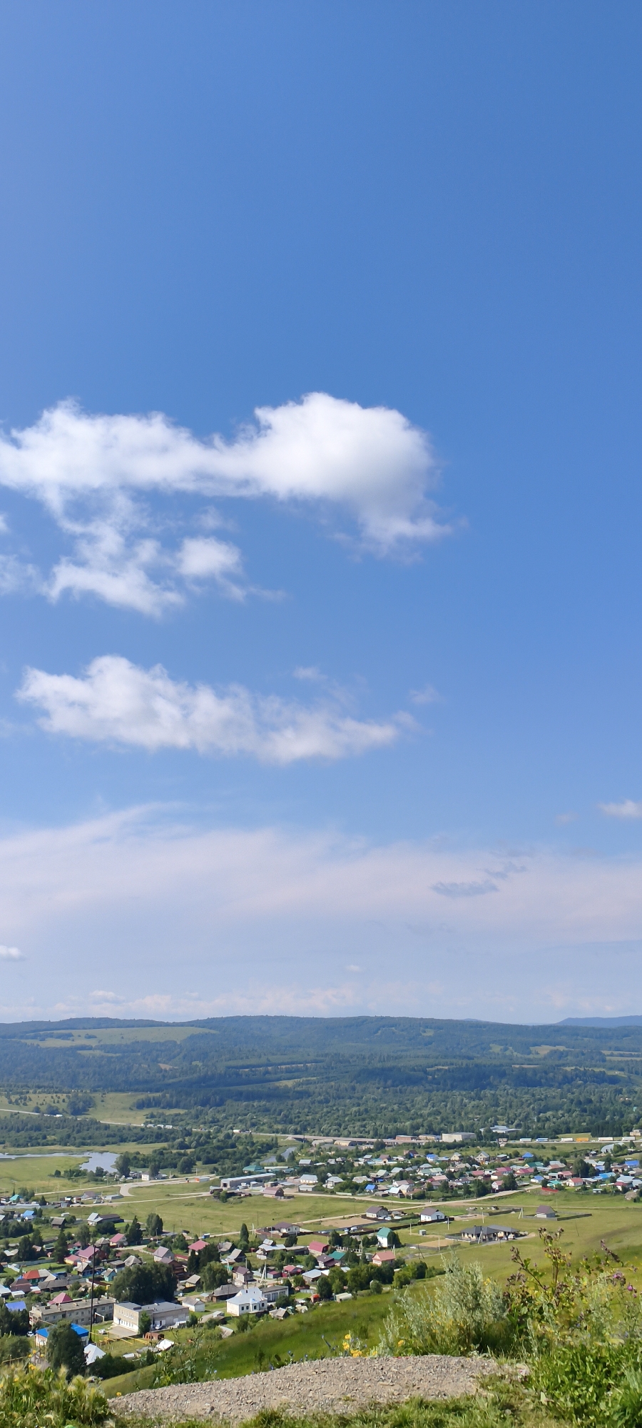 The image shows an aerial view of the city of Sim, Russia, on a clear, sunny day. The sky is a vibrant blue with scattered white clouds. Below the sky, a range of rolling, tree-covered hills forms the horizon. In the mid-ground, the city of Sim spreads out, characterized by a dense collection of houses with colorful roofs, interspersed with green spaces and trees. Roads and pathways wind through the residential areas. A body of water, possibly a river or lake, is visible to the left of the city, reflecting the daylight. The foreground consists of a grassy slope covered with small rocks and sparse vegetation, suggesting the viewpoint is from a higher elevation looking down upon the city. There are no people or animals visible in the frame. The scene depicts a typical rural or small-town settlement nestled within a natural landscape.