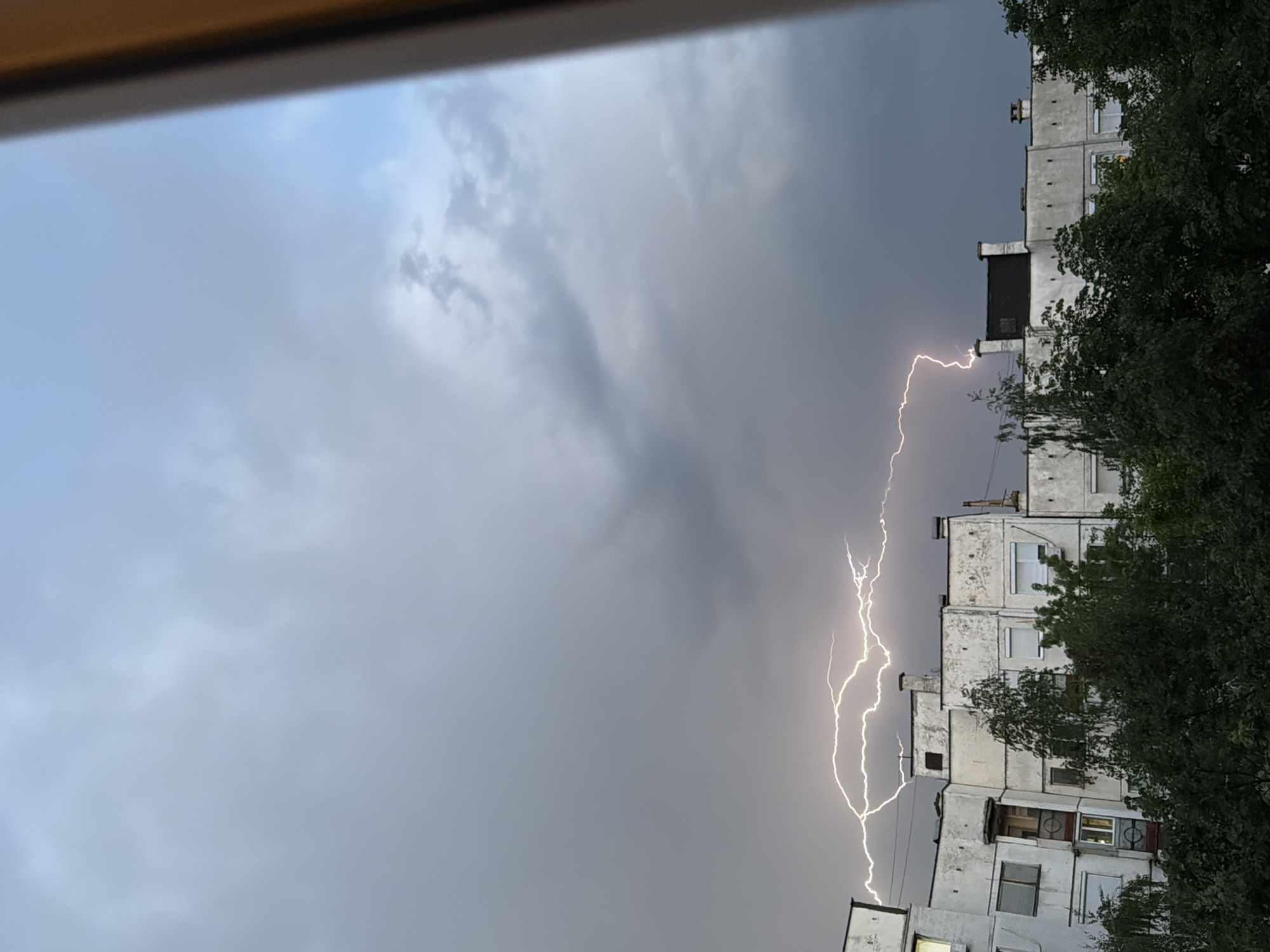 A view through a window captures a lightning strike near a tall building in Pleven, Bulgaria. The sky is overcast and dark, with dense gray clouds filling most of the frame. A bright, forked lightning bolt is prominently visible, descending vertically alongside the side of the building. The building is a multi-story structure with multiple windows, appearing to be an apartment complex. Some windows are dark, while others show signs of interior lighting. Parts of the building are partially obscured by trees with dense, green foliage. The window frame through which the scene is viewed is visible at the top of the frame.