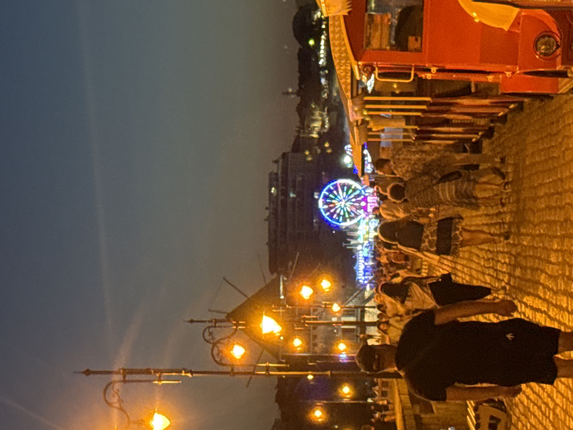 The image depicts a street scene at dusk in Nesebar, Bulgaria, likely on the peninsula where the old town is located. In the foreground, a man wearing a black t-shirt and shorts walks away from the camera. Behind him, a group of people are walking along a cobblestone path illuminated by street lamps. To the right, a red tourist train with several carriages is parked. In the background, a Ferris wheel glows with colorful lights, and beyond that, buildings are visible against the darkening sky. The atmosphere suggests a lively evening, possibly a tourist area given the presence of the train and Ferris wheel.