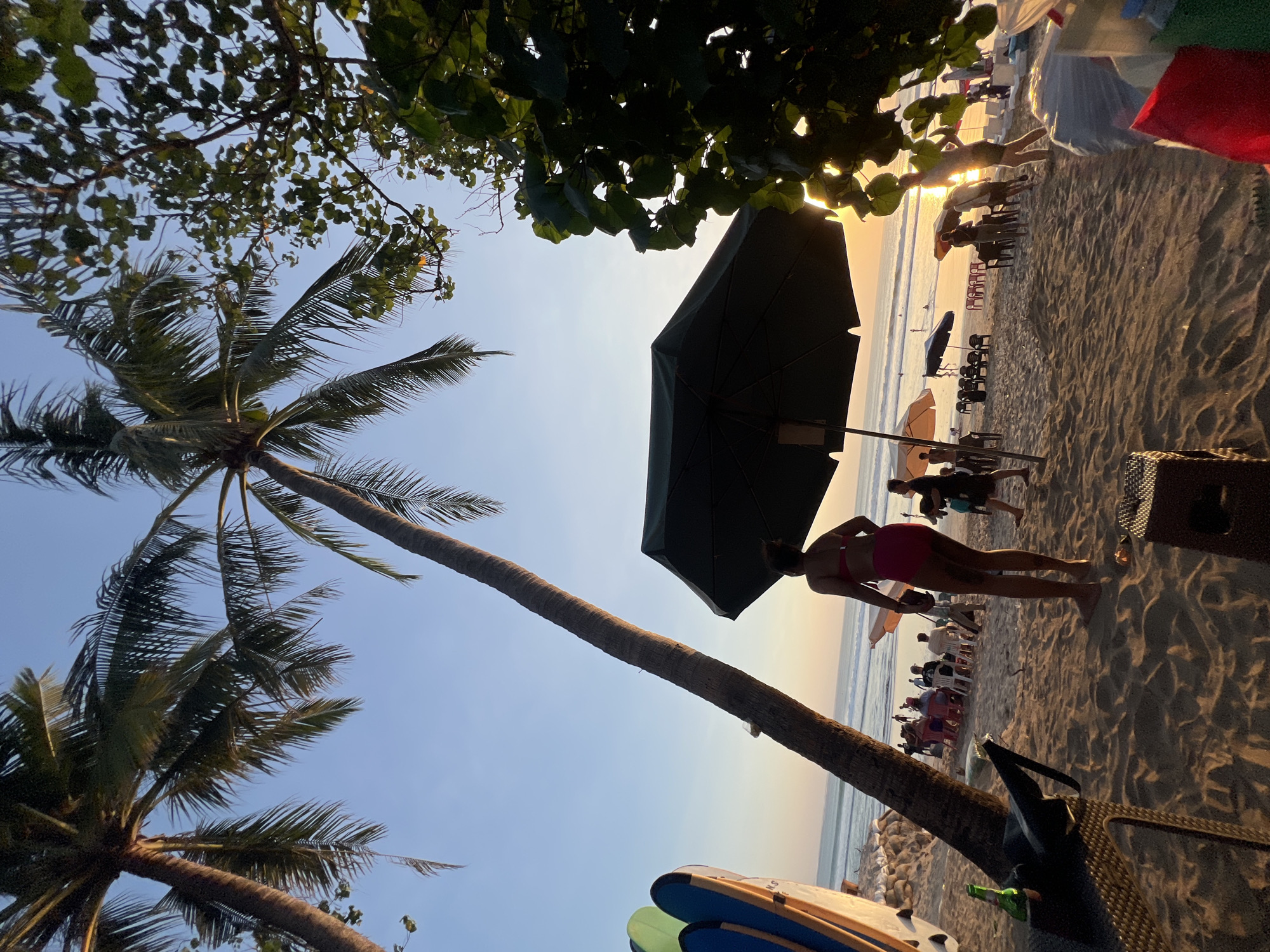 This is a photograph taken at sunset on Kuta Beach, Bali, Indonesia. The foreground is dominated by sand dunes and a large, dark green umbrella providing shade. To the left, surfboards lean against a textured surface, and a bottle of beer rests on a woven table. In the middle ground, a woman in a pink bikini stands facing away from the camera, looking out at the ocean. Several beach chairs and umbrellas are scattered across the sand, occupied by various people relaxing or conversing. The ocean stretches to the horizon, with gentle waves rolling in and a few individuals wading in the water. Palm trees, silhouetted against the sky, frame the scene on the left and center. The sky is a gradient of pale blue and warm orange, indicative of the setting sun. The overall atmosphere is one of a tranquil beach evening.