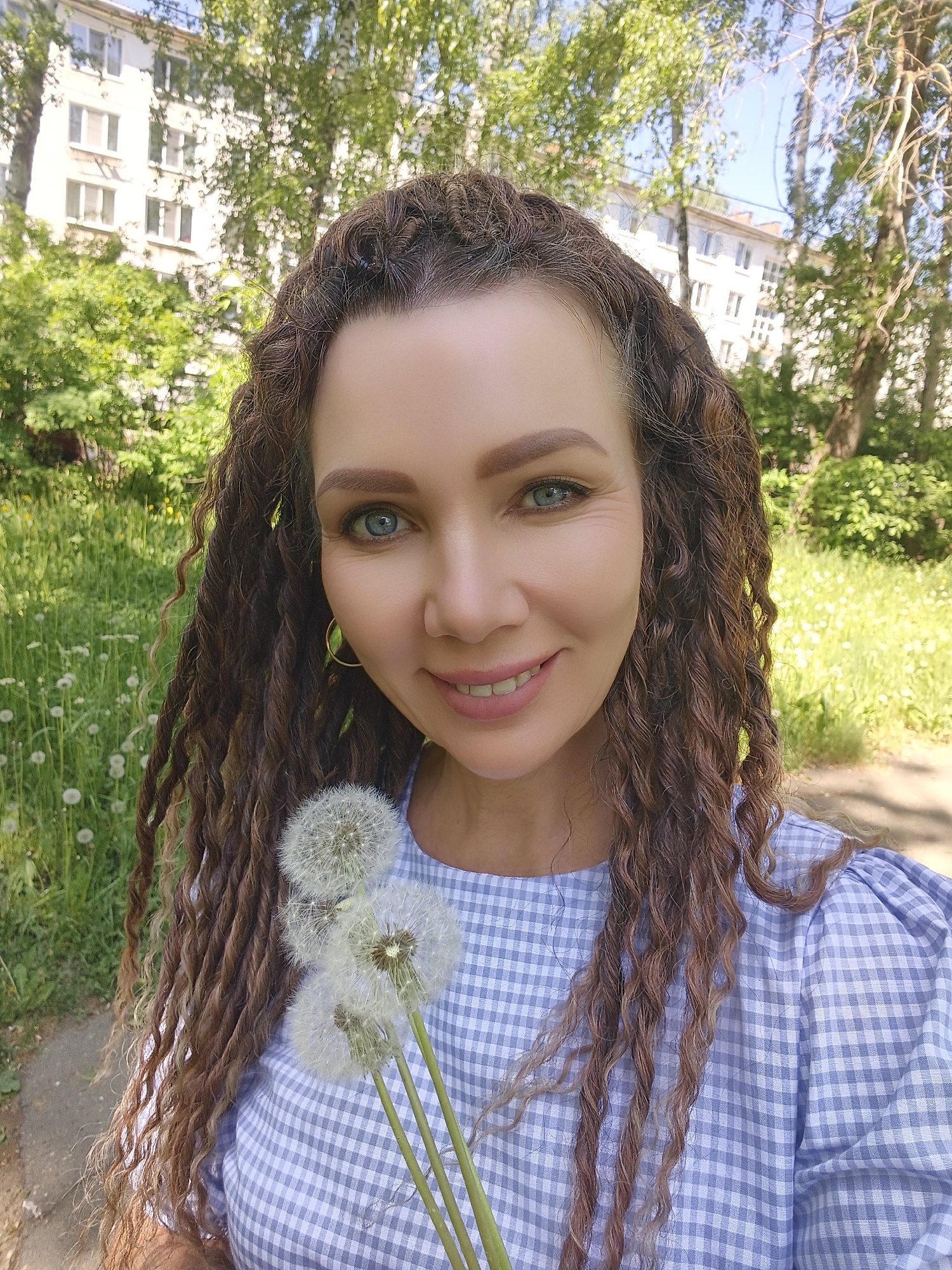 A woman with long brown curly hair, fair skin, and blue eyes smiles while holding a small bouquet of dandelions in a park. She wears a blue and white checkered blouse with puffed sleeves. The background includes green trees, a patch of dandelions, and a white building. The location is a park in a city.