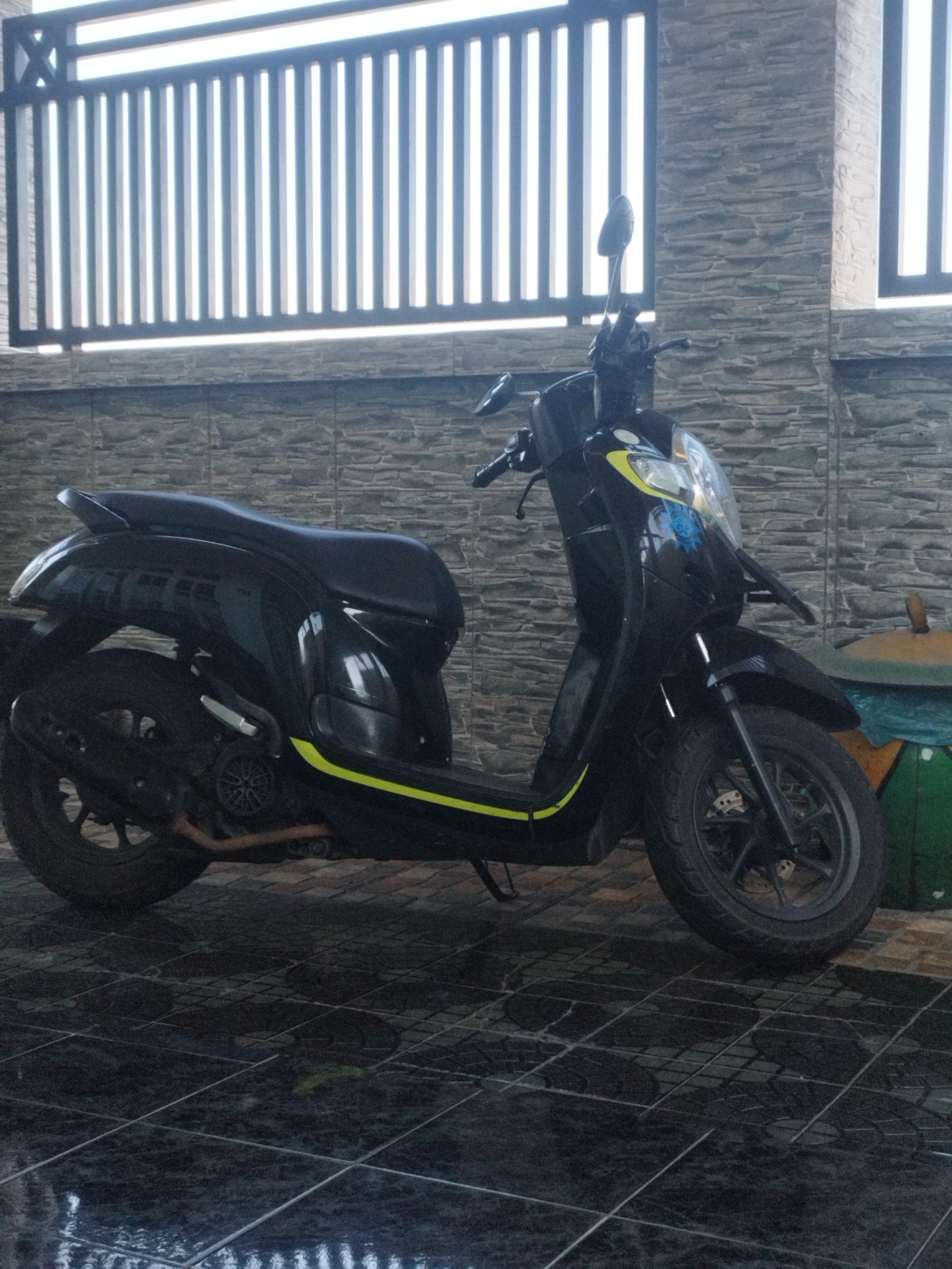 An all-black scooter with yellow accents is parked on a wet, black tiled floor in front of a stone-tiled wall. Above the wall is a dark metal fence with vertical slats. The scooter has a black seat, black wheels with visible brake discs, and a black exhaust pipe. A yellow stripe runs along the side of the scooter. The scooter has a headlight with a blue decal on it. To the right of the scooter, partially visible, is a large, round, olive-green container with a yellow lid. The overall scene appears to be an outdoor covered area, possibly a carport or patio, in Sumenep, Indonesia.