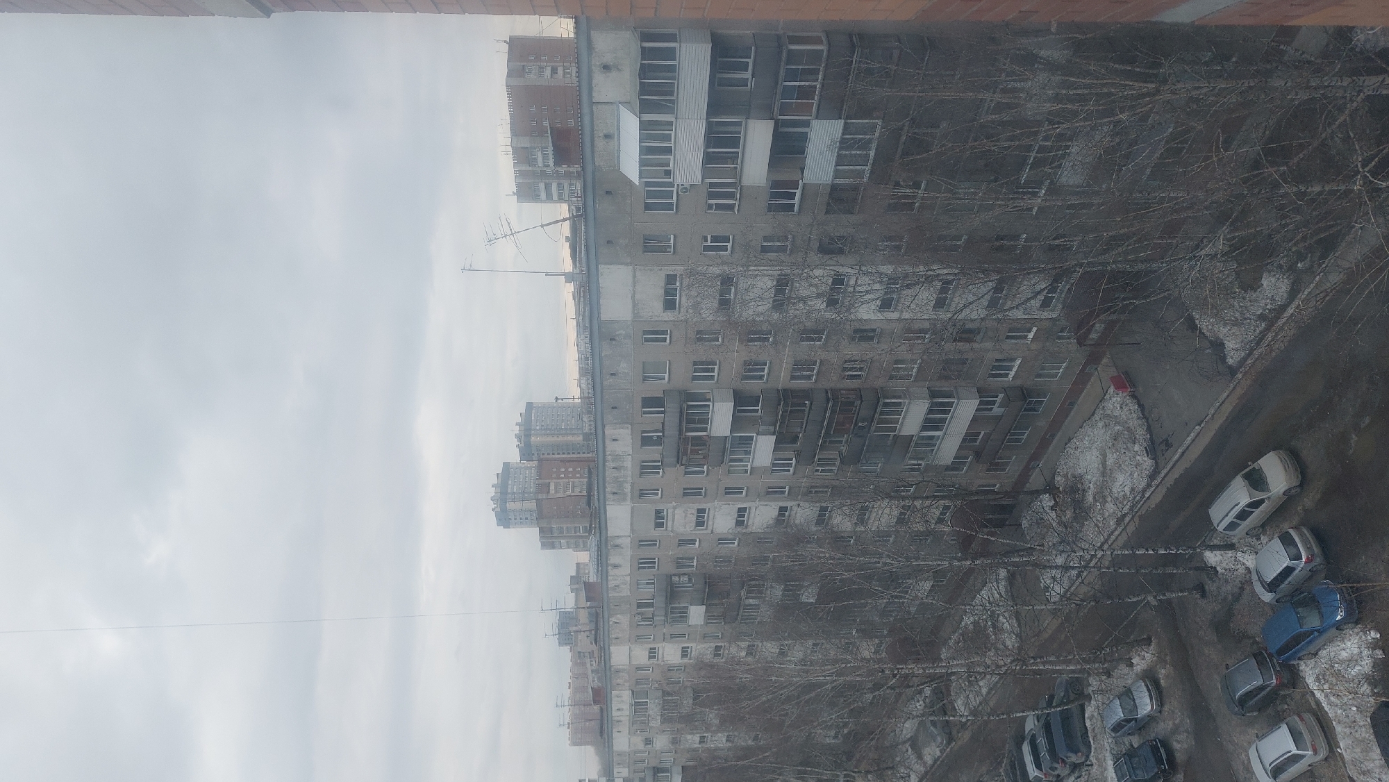 The image shows a view from a high vantage point in Novosibirsk, Russia, overlooking a residential area characterized by Soviet-era apartment blocks. These buildings are multi-story, concrete structures with repetitive window patterns and balconies. Bare trees line the street, and some snow is visible on the ground. Parked cars are scattered along the roadside below. The sky is overcast, suggesting a cold, possibly wintery day.