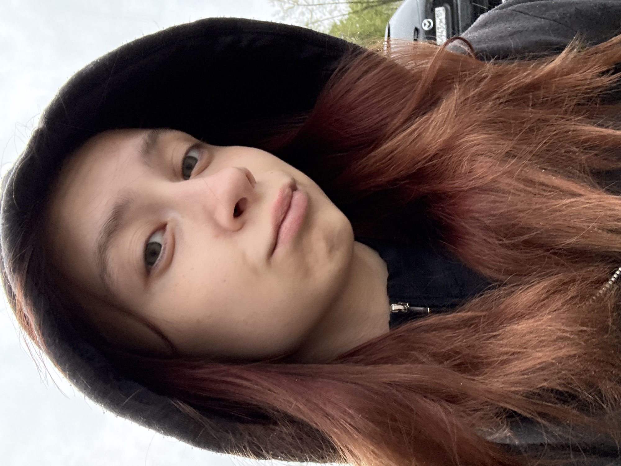 A close-up, low-angle selfie features a young person with auburn hair and fair skin. They are wearing a dark hooded sweatshirt with the hood up. The person's face is visible, showing their eyes, nose, and mouth with a slight frown. The background is blurred and indistinct, but hints of greenery and a vehicle suggest an outdoor setting, possibly a street. The image could be taken at the city of Chaikovsky, Russia, however, the location is unclear from visual elements.