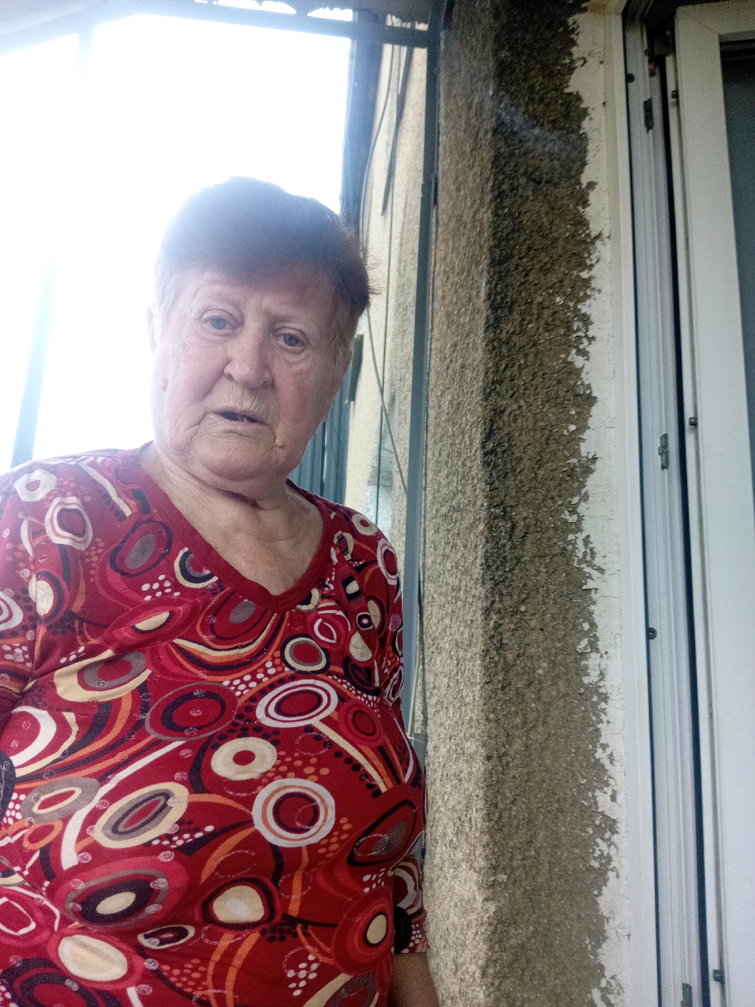 An elderly woman with short, reddish-brown hair and fair skin is standing in front of a building. She is wearing a red, v-neck shirt with a colorful, abstract pattern. Her mouth is slightly open, and she is looking directly at the camera. To the right of the woman, a section of the building's exterior wall is visible. The wall appears to be made of concrete or stucco and has a rough texture with patches of dark discoloration, suggesting water damage or mold. A white window frame is also visible next to the wall. The background is largely out of focus, with a bright, blown-out area suggesting strong light from a window or the sky. The scene is likely outdoors, possibly on a balcony or near an entrance. The image appears to have been taken in Tiachiv, Ukraine, based on the provided context.