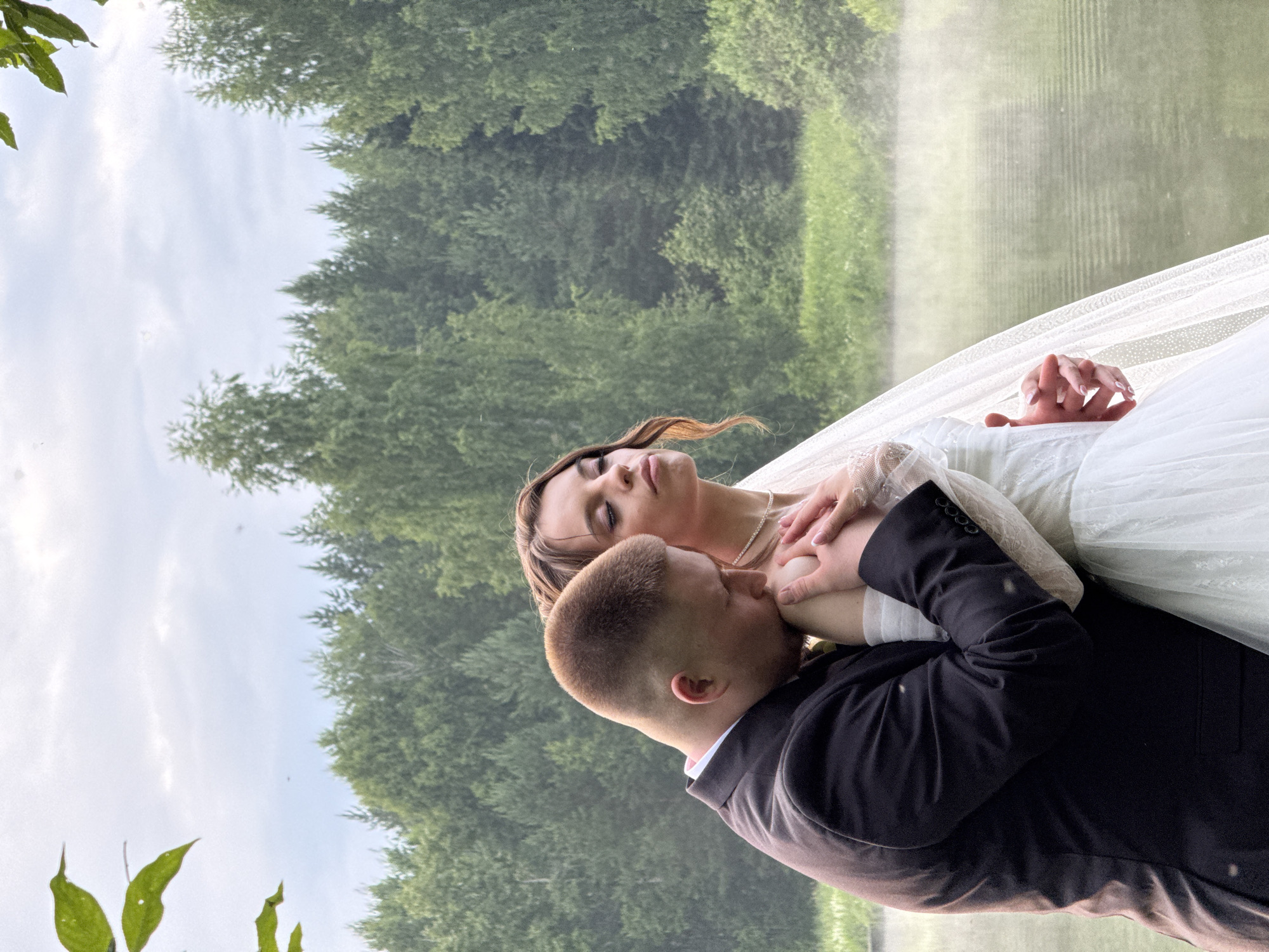 A wedding portrait features a bride and groom in an embrace. The bride, wearing a white wedding dress and veil, is resting her head on the groom's shoulder. The groom, dressed in a black suit with a white shirt, is kissing her shoulder. Their hands are intertwined, with the bride's ring visible on her left hand. The couple appears to be in a natural setting, with a body of water and lush green trees in the background. The lighting suggests it is daytime, possibly overcast given the soft light. The mood is intimate and romantic. The image is oriented vertically, with the sky and treetops at the top and bottom of the frame. The location is Votkinsk, Russia. There is no visible text in the image.