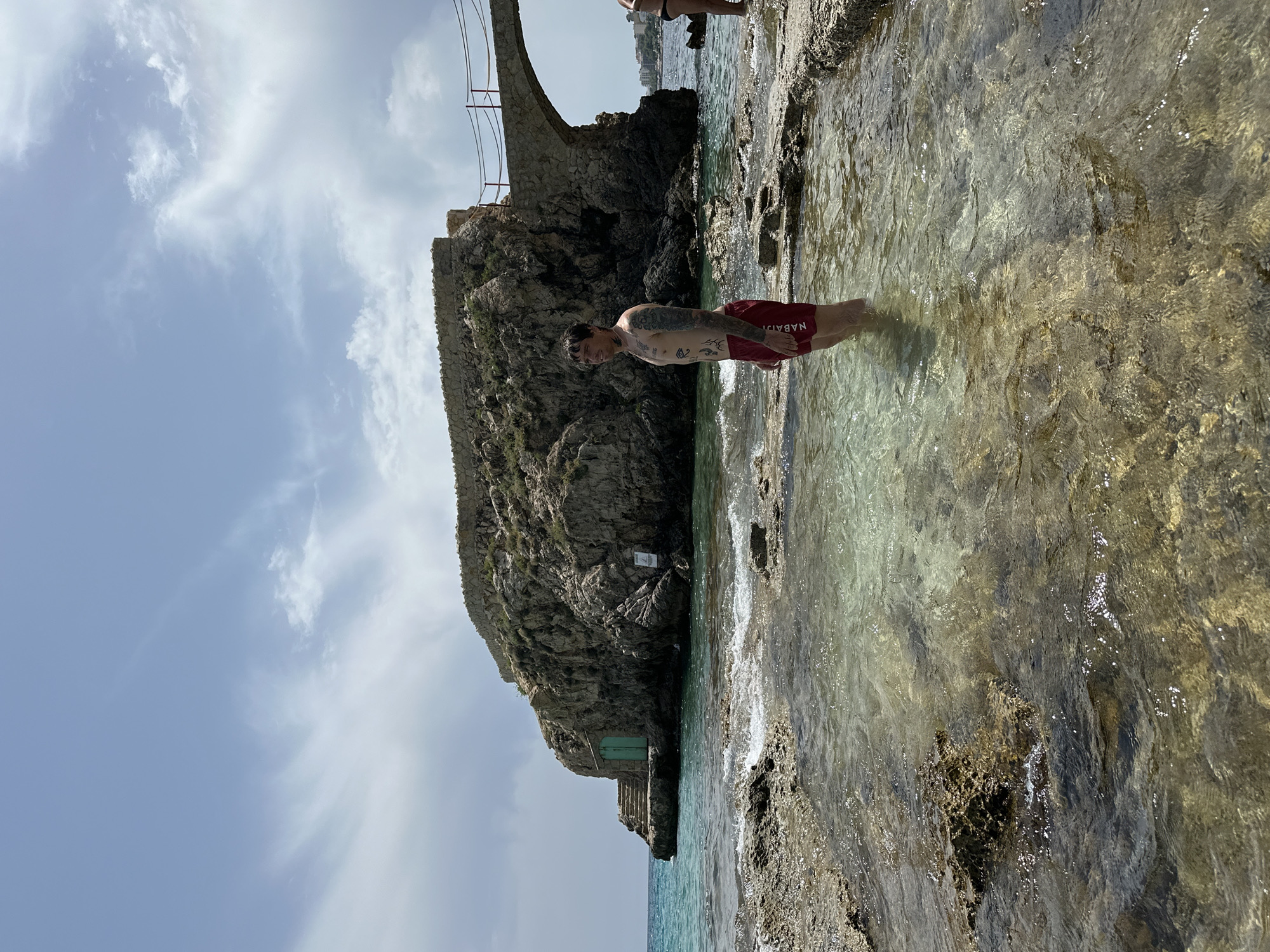 In Sant Agustí, Spain, a man with extensive tattoos stands in shallow, clear water near a rocky coastline. He is wearing red swimming trunks. Behind him, a large rocky outcrop juts out of the sea, connected by a stone arch bridge to another rocky formation. A small, green door is visible on the side of the rocky outcrop, accessible by a set of stone steps. The water is calm, with small waves lapping against the rocks in the foreground. The sky above is partly cloudy, with a bright blue sky peeking through.