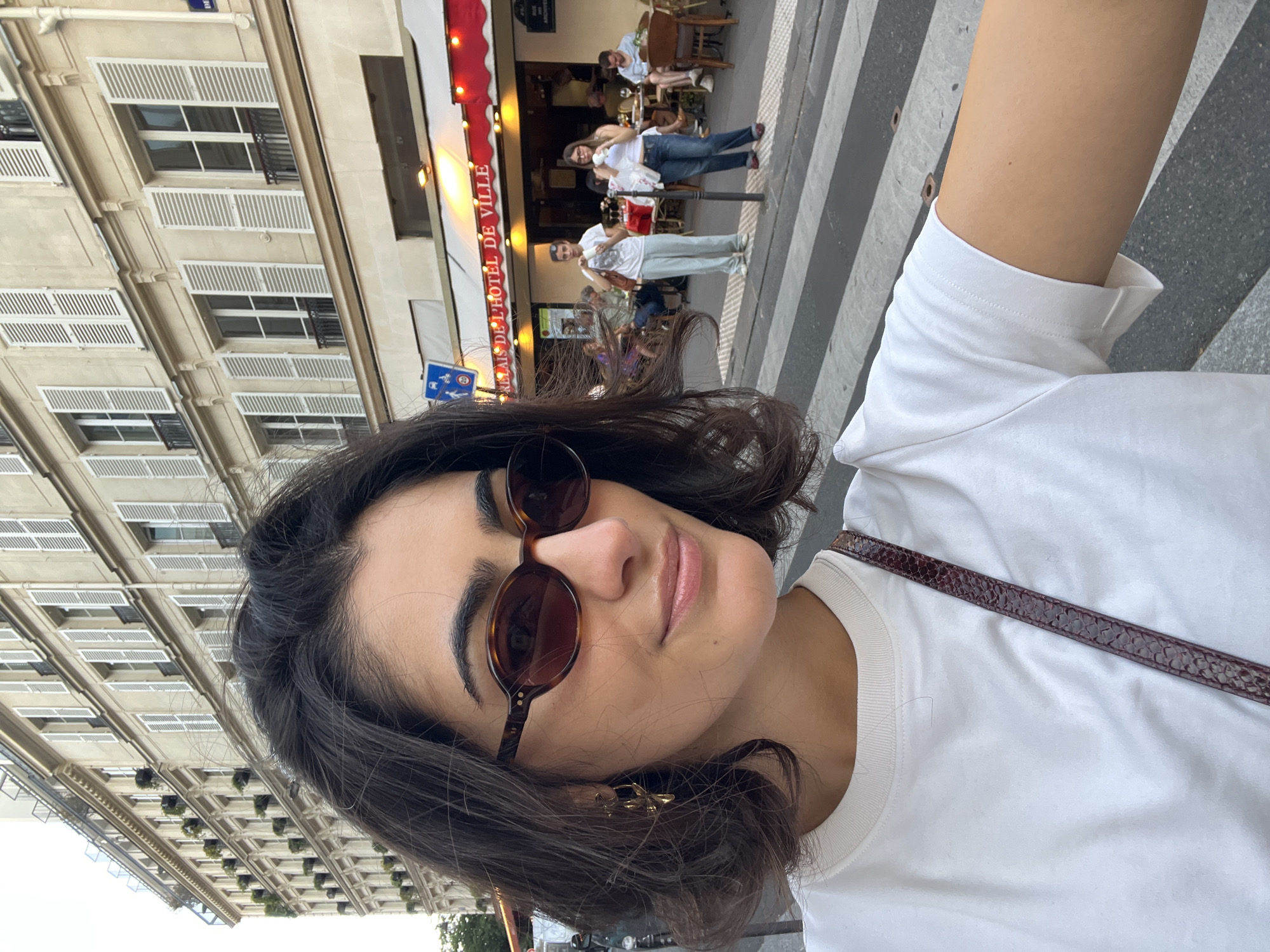 The image appears to be a selfie taken in Paris, France, with the camera held at an angle. A young woman with dark hair and sunglasses is in the foreground, wearing a white T-shirt. A patterned strap crosses her chest. Behind her, a Parisian street scene is visible, featuring buildings with white shuttered windows and a restaurant or cafe with an awning that reads "PALAIS DE L'HOTEL DE VILLE." People are seated and standing outside the establishment. A crosswalk stretches across the foreground of the street. A blue sign with white icons is attached to the building, presumably a street sign.