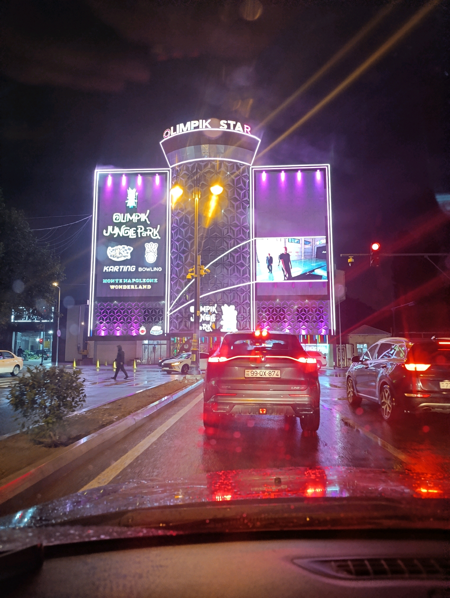 The image depicts a nighttime street scene in Baku, Azerbaijan, likely in the vicinity of the "Olimpik Star" entertainment complex. A multi-story building with prominent neon signage reading "OLIMPIK STAR" is illuminated, with the words "OLIMPIK JUNGE PARK" also visible. Below this, "KARTING BOWLING" and "MONTE NAPOLEONE WONDERLAND" are advertised. The building's facade is decorated with intricate geometric patterns and vertical lines of bright white lights, interspersed with purple lighting. Two yellow spotlights are projected upwards from the building.
The street is wet, indicating recent rain, and the wet asphalt reflects the colorful lights of the complex and vehicle brake lights. Several cars are on the road. In the foreground, the back of a dark-colored SUV is visible, with the license plate reading "99-QX-874". To its right, another car, a silver SUV, is also stopped. In the background, a pedestrian is walking on the sidewalk. Traffic lights are visible, with one red light illuminated. The sky is dark, with some diffused lighting and what appear to be light beams or reflections from the building's lights. The overall atmosphere is one of a brightly lit urban entertainment area at night, with wet weather conditions.