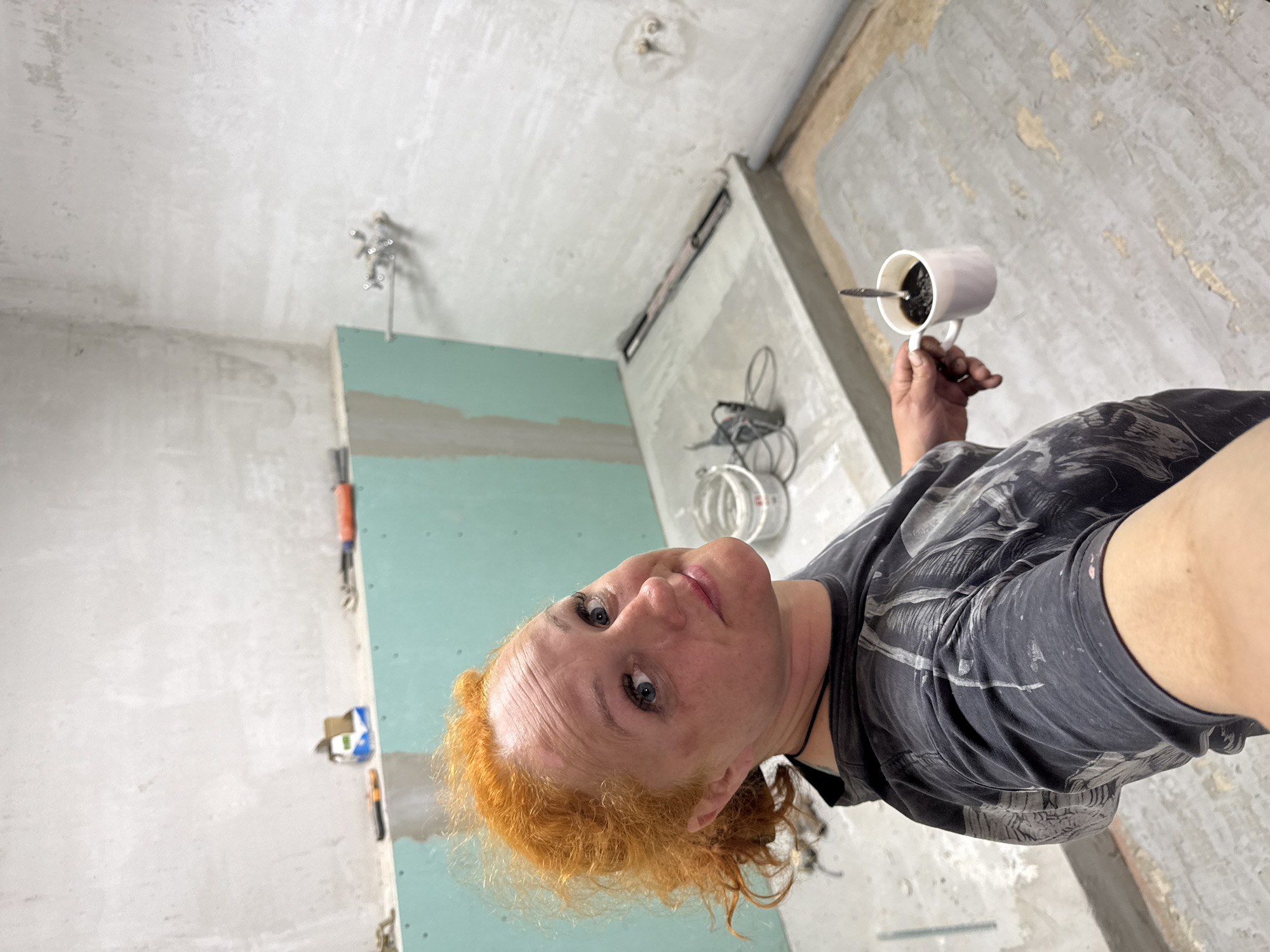 The image shows a person indoors, presumably in the city of Chaikovsky, Russia, given the prompt context. The individual, a woman with red hair, is holding a white mug filled with a dark liquid, possibly coffee, with a spoon in it. She is wearing a dark-colored t-shirt that appears to have paint or other stains. The background suggests an ongoing renovation or construction project. The walls are unfinished, with some sections covered in green drywall. Tools are visible hanging on the walls, and electrical wiring is exposed. The ceiling is also unfinished, with what appear to be fixture connections exposed.