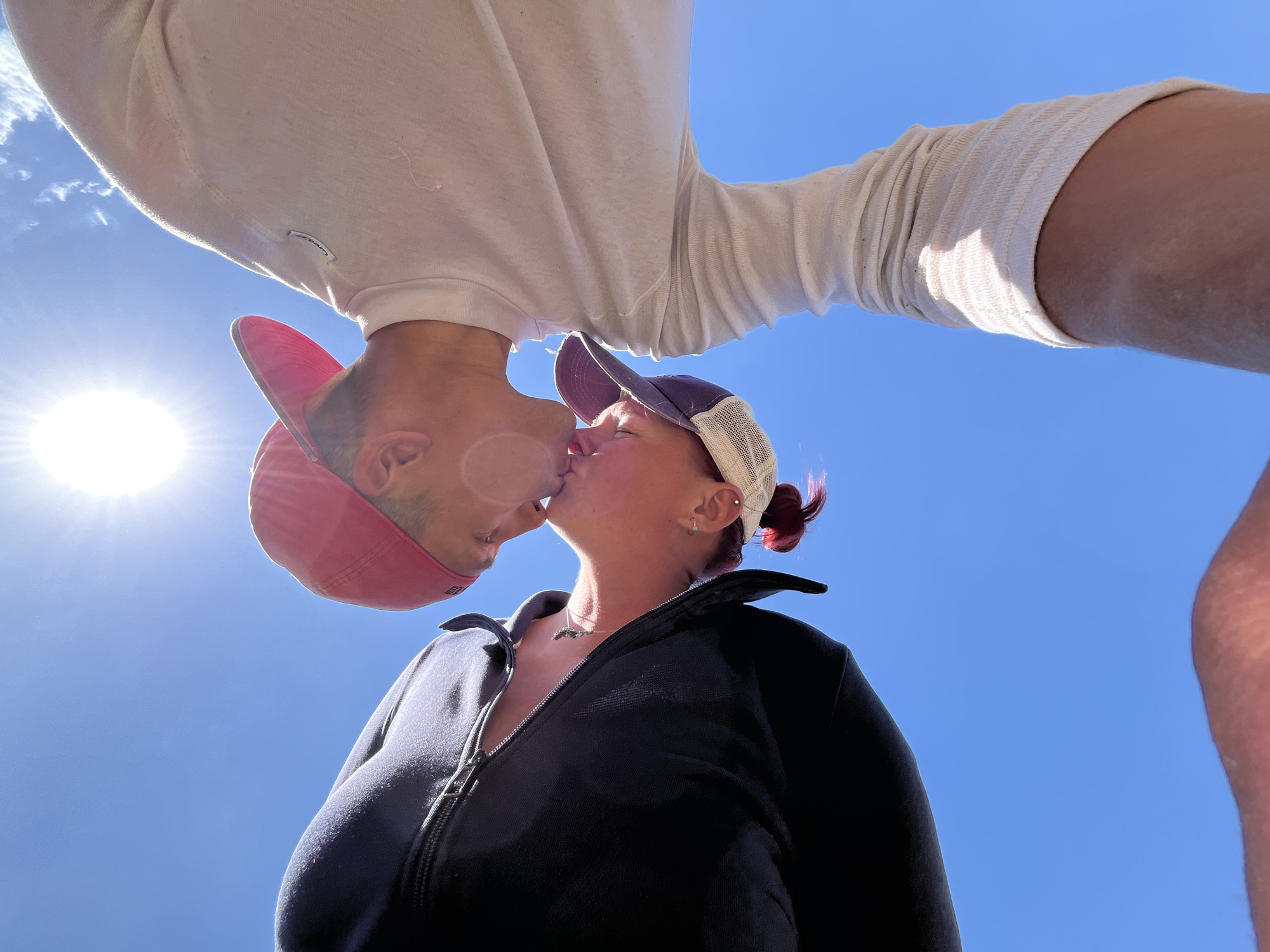 A low-angle photograph depicts two people kissing against a bright blue sky with a sun visible in the upper left. The person on the left wears a red cap and a white long-sleeved shirt. The person on the right wears a black zip-up jacket and a dark blue and white trucker hat. The location cannot be determined from the image, so the specification of Chaikovsky, Russia cannot be confirmed.