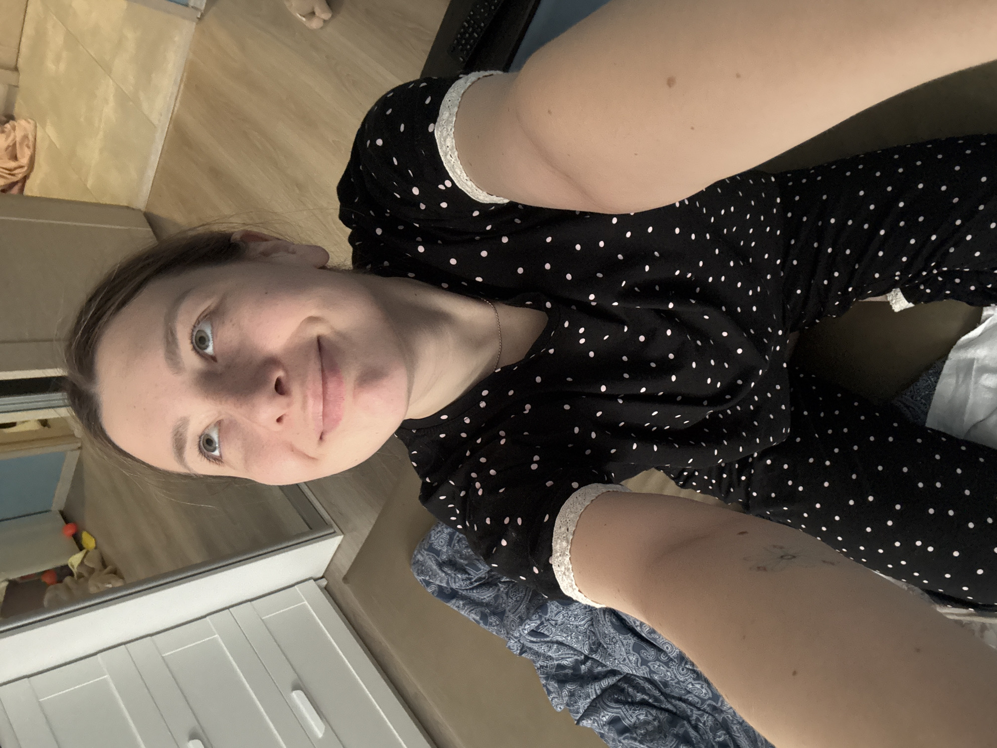 A woman is taking a selfie. She is wearing a black and white polka dot shirt with white lace trim on the sleeves. She is looking directly at the camera with a slight smile. The background includes a white cabinet with drawers, a wooden floor, and a blue patterned blanket or fabric. The location is not identifiable as Moscow, Russia based on the provided image.
