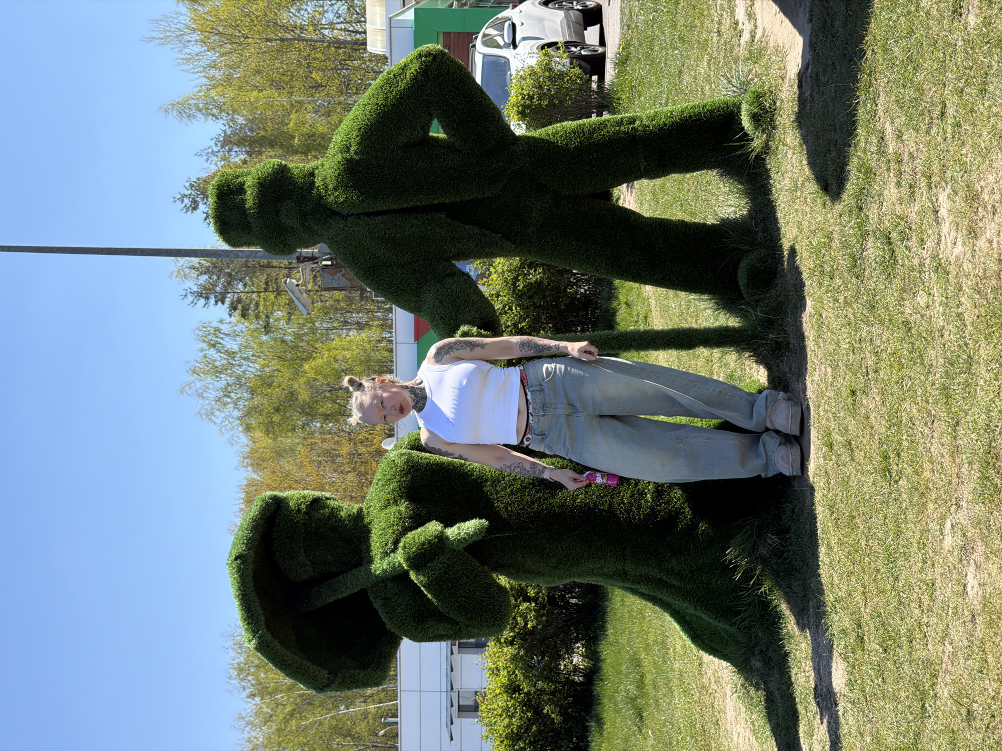A young adult woman with tattooed arms and blonde hair in a bun is standing between two green topiary figures on a grassy lawn. The woman is wearing a white tank top, blue jeans, and tan shoes, and is holding a small pink and purple object in her right hand. One topiary figure in front of her is holding what appears to be an umbrella, and the other behind her is in a walking or striding position. Behind the figures and woman, trees, a white car, and a low-rise building are visible. The location is the city of Крошынскі сельскі Савет, Belarus. The sky is clear and blue.