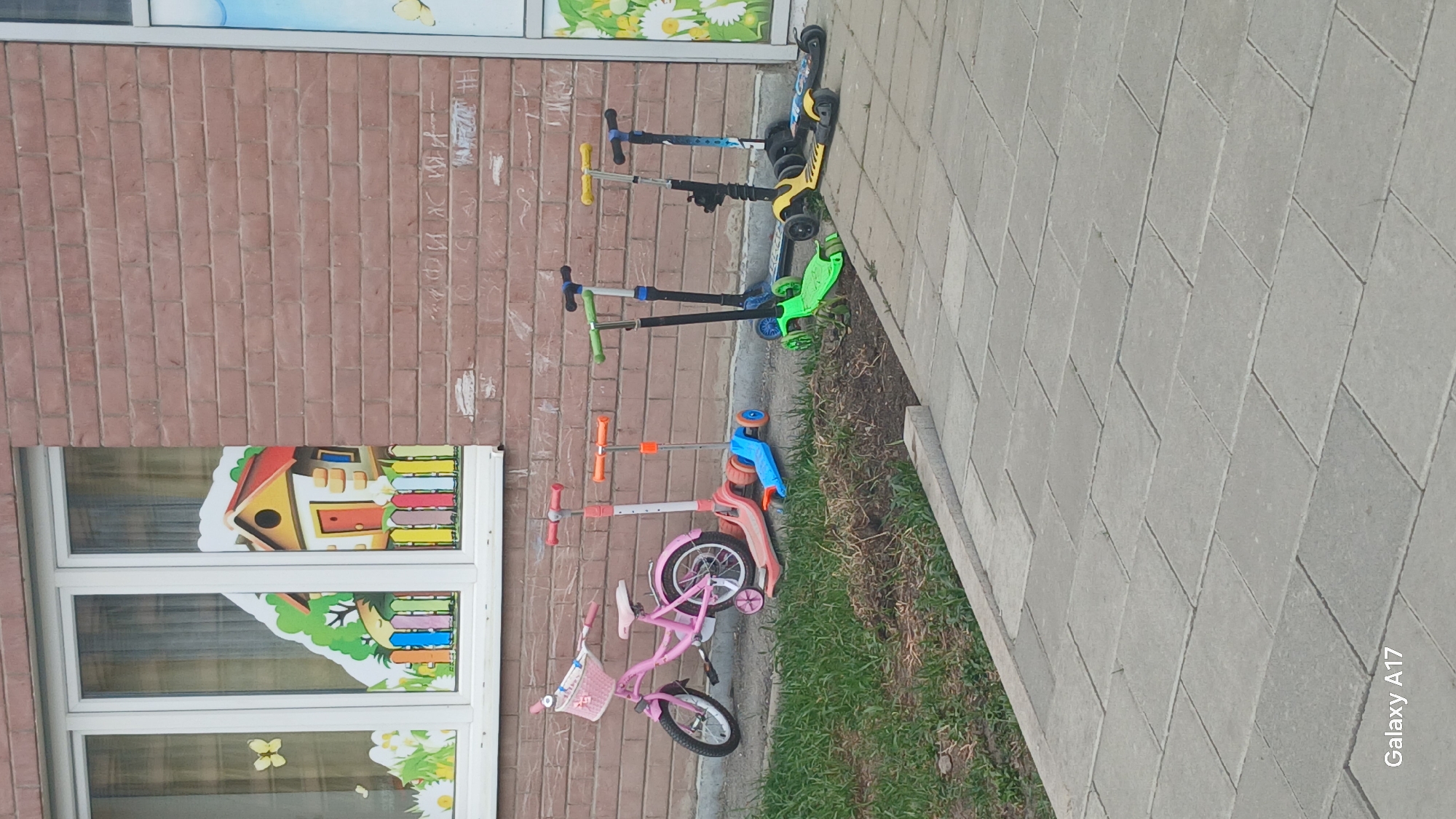 The image shows a collection of children's scooters and a small bicycle leaning against a brick wall. The wall has a window with colorful stickers depicting a house and other images associated with children. The scooters are various colors and styles. The bicycle is pink with a basket. The background includes a paved area and some grass. The location is Yekaterinburg, Russia.